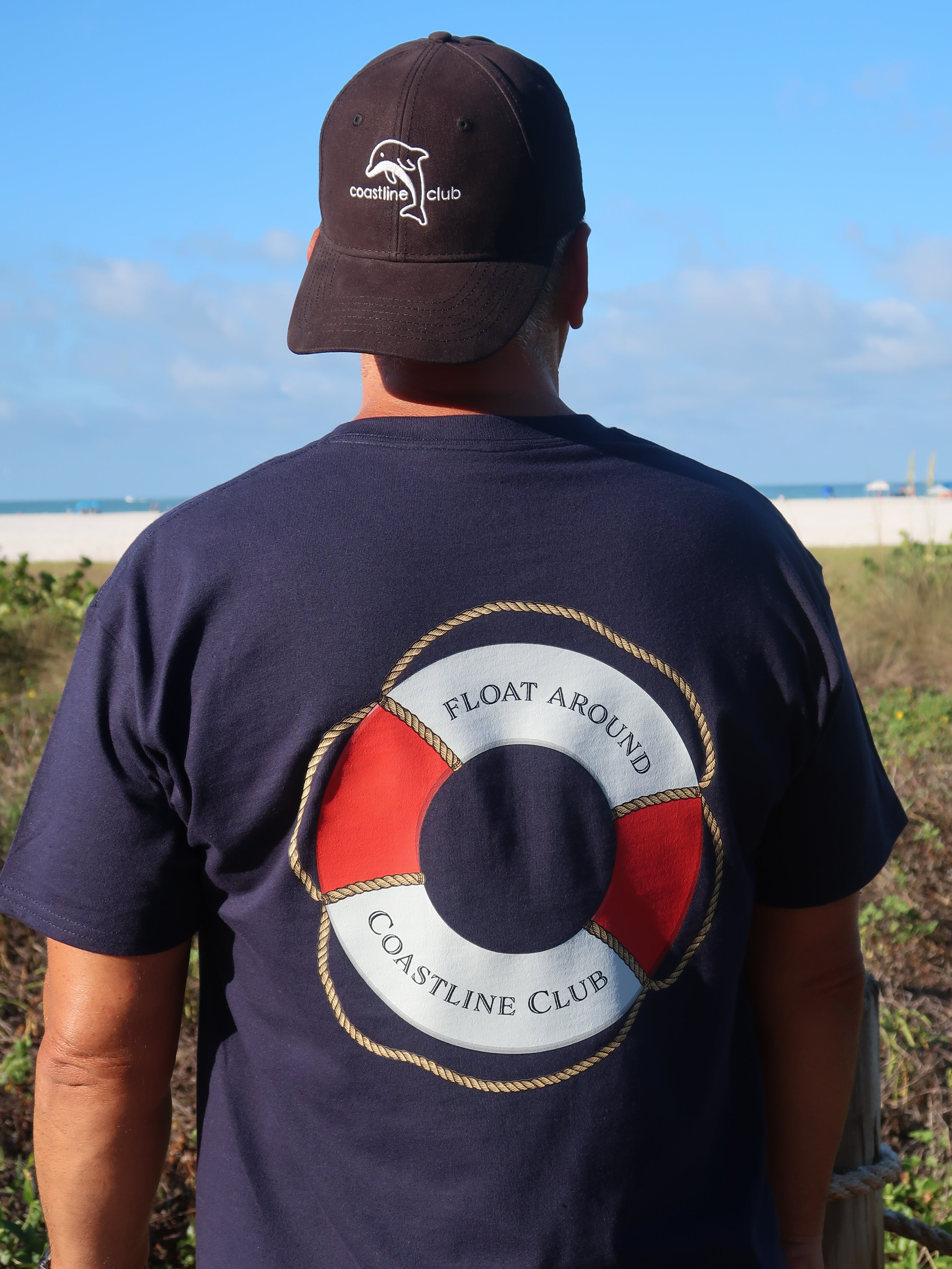 Man wearing a navy blue t-shirt with a life buoy design and 'Coastline Club' text, standing at a beach.