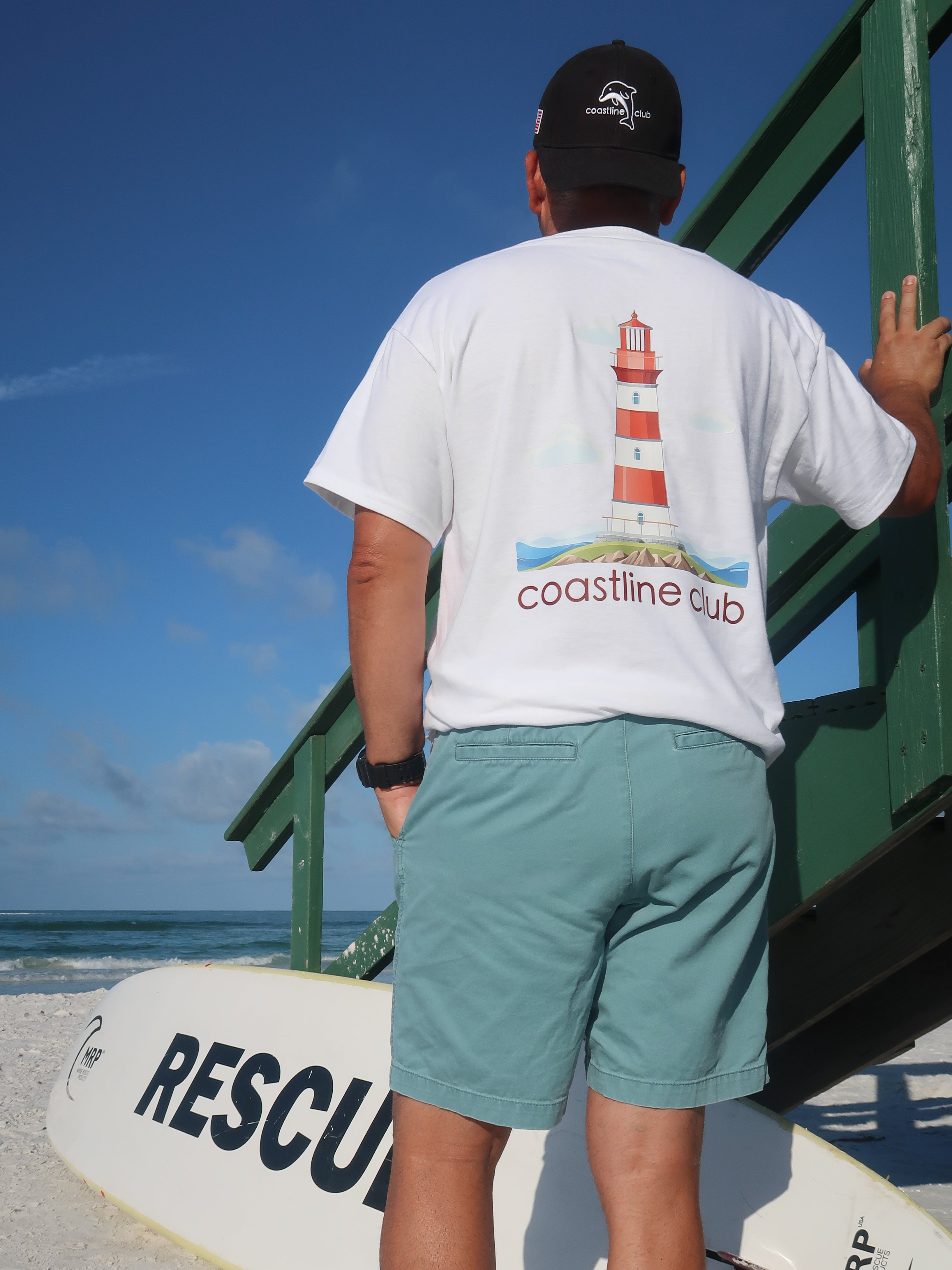 Person wearing a white t-shirt with a lighthouse design and 'coastline club' text on a beach.