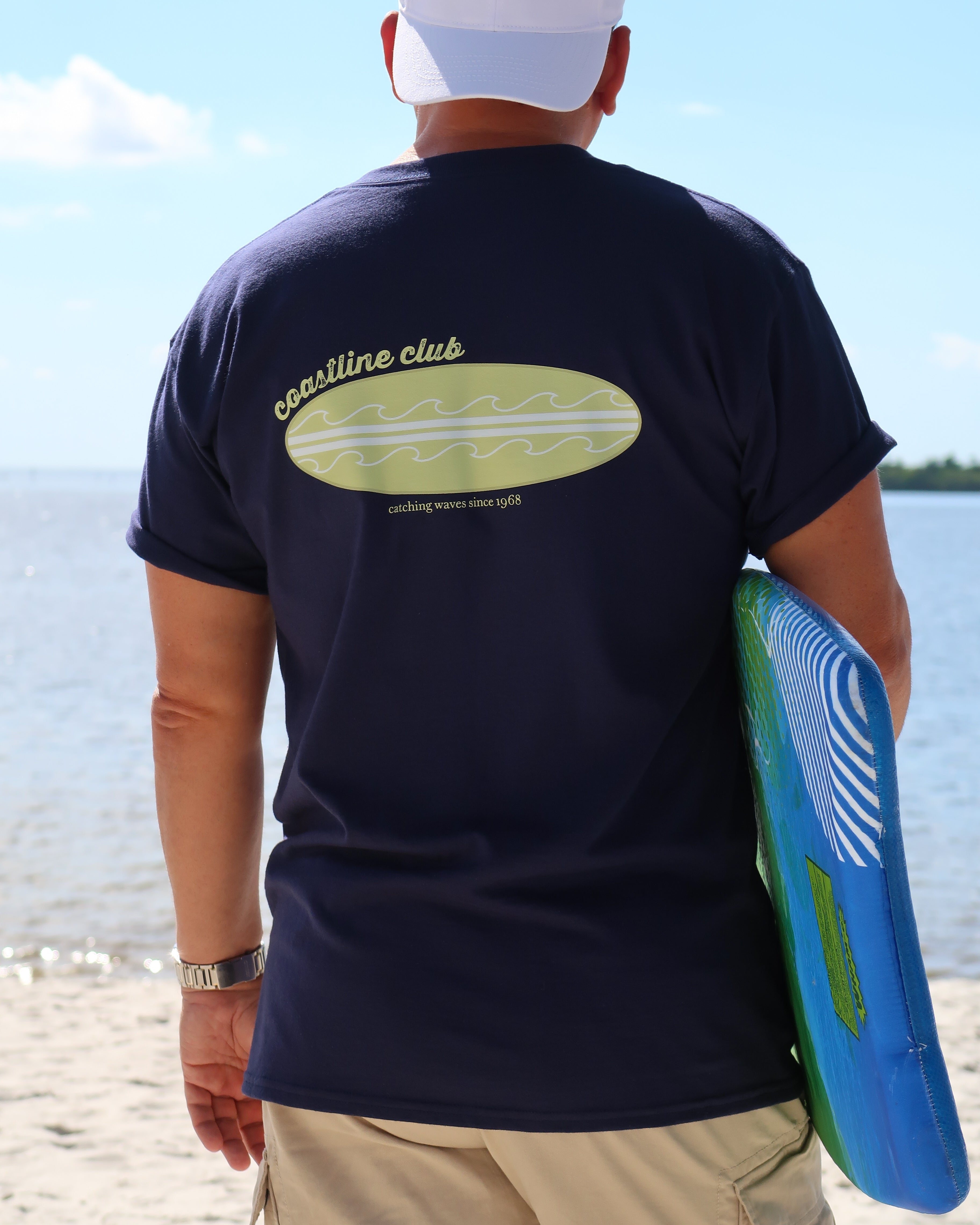 Person wearing a navy blue t-shirt with a surfboard graphic on a beach
