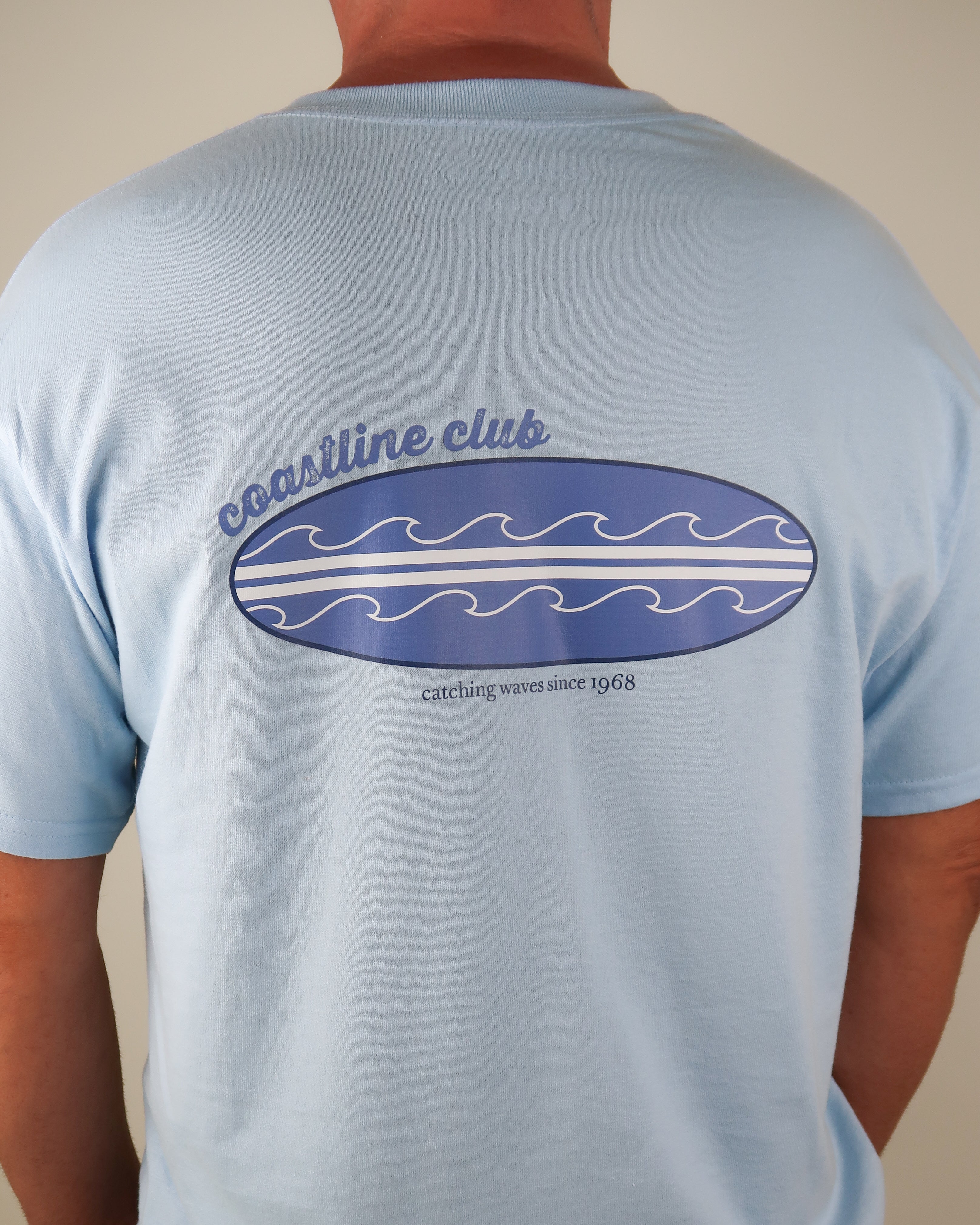 Light blue t-shirt with 'coastline club' surfboard on the back, worn by a person.
