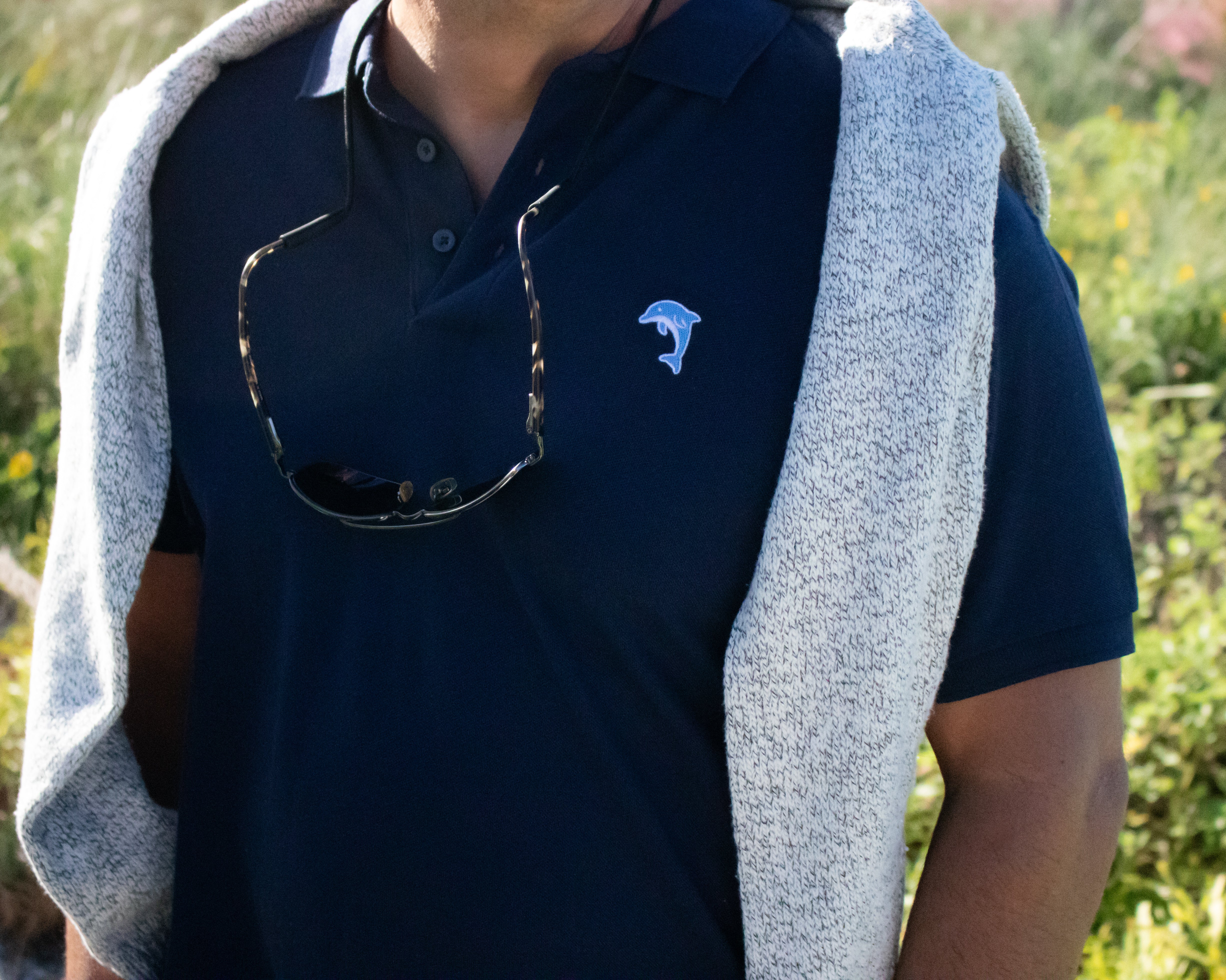 Person wearing a navy polo shirt with a logo and a gray sweater outdoors.