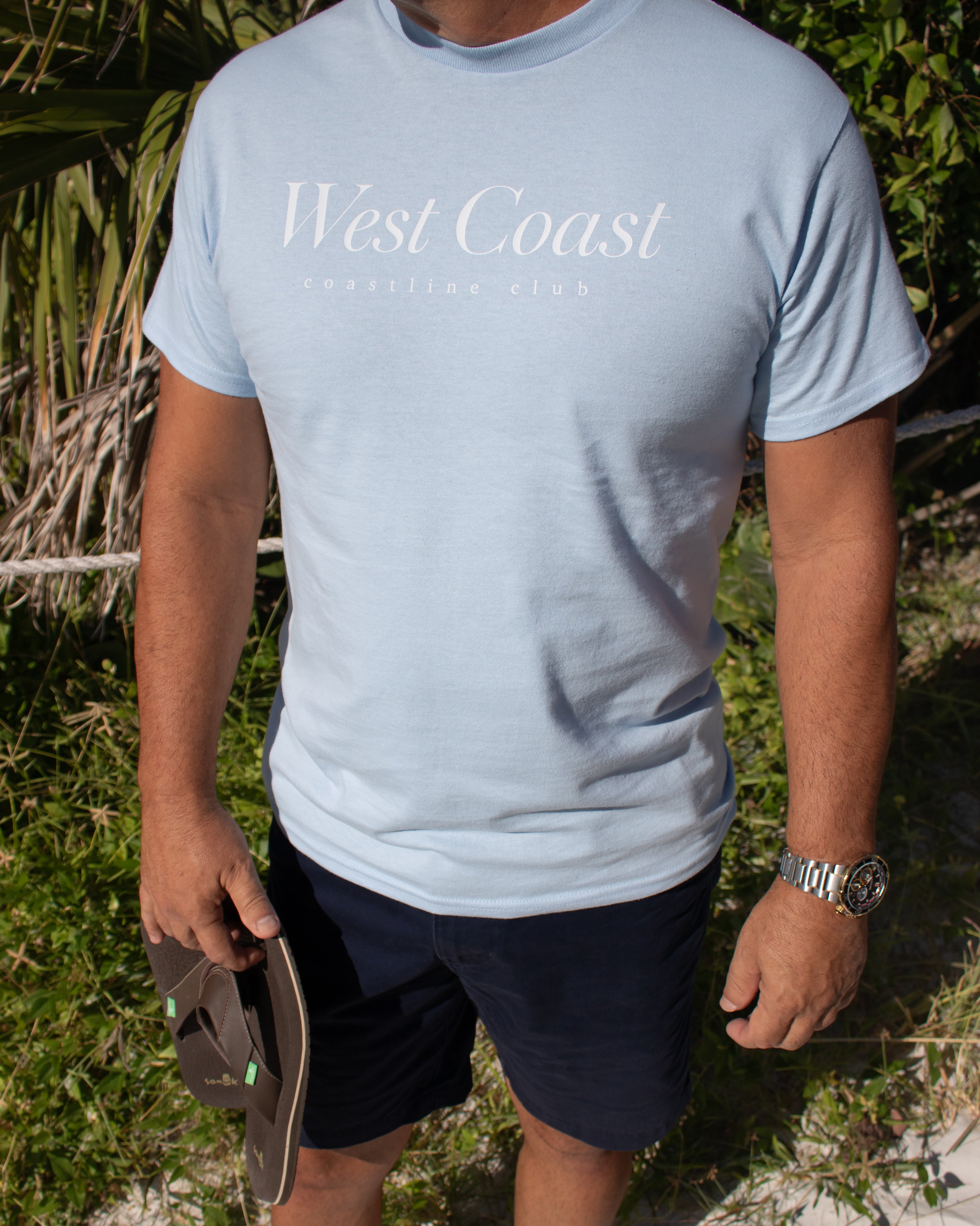 Person wearing a light blue 'West Coast' t-shirt outdoors.