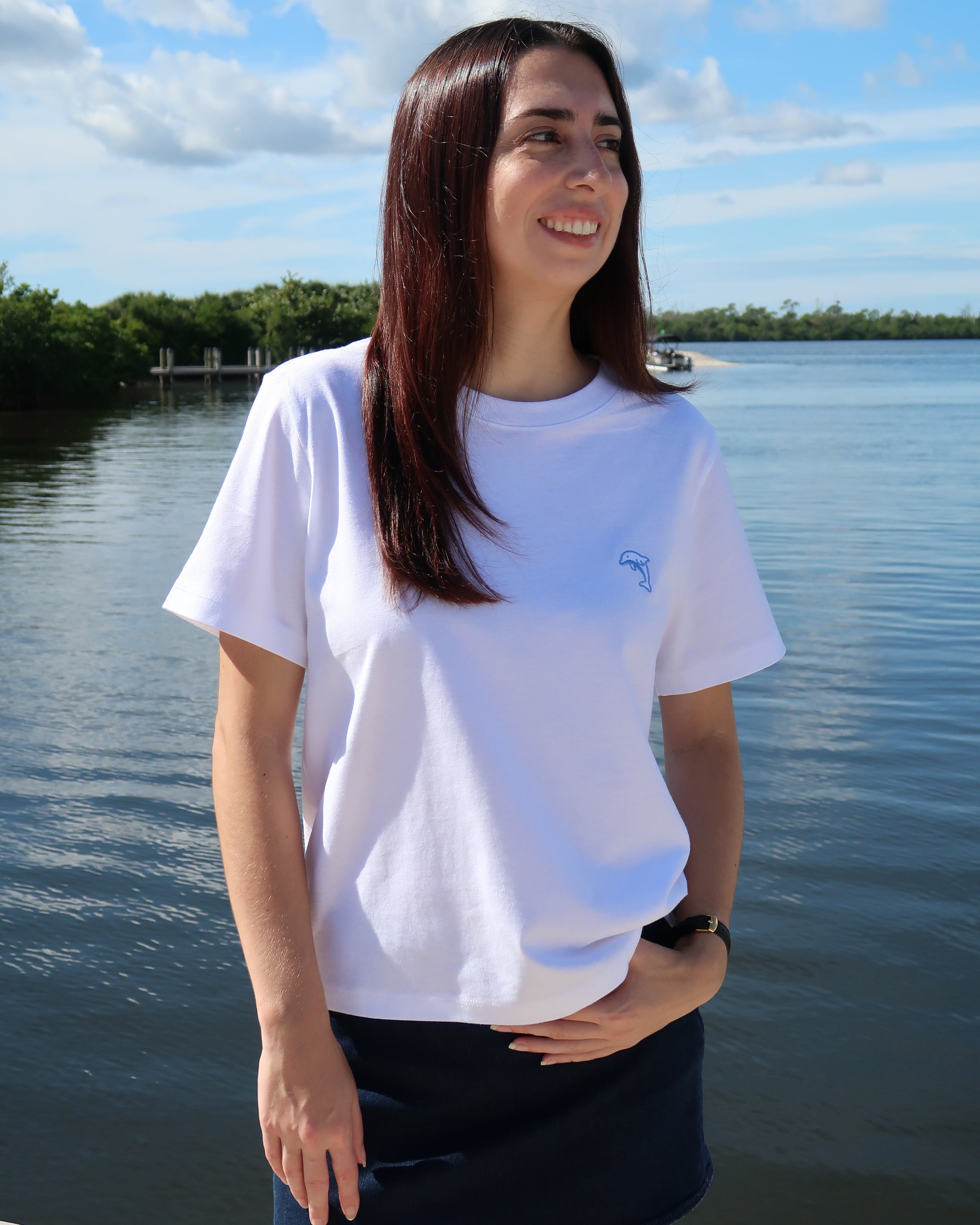 Woman wearing a white t-shirt with a dolphin logo standing by a body of water.