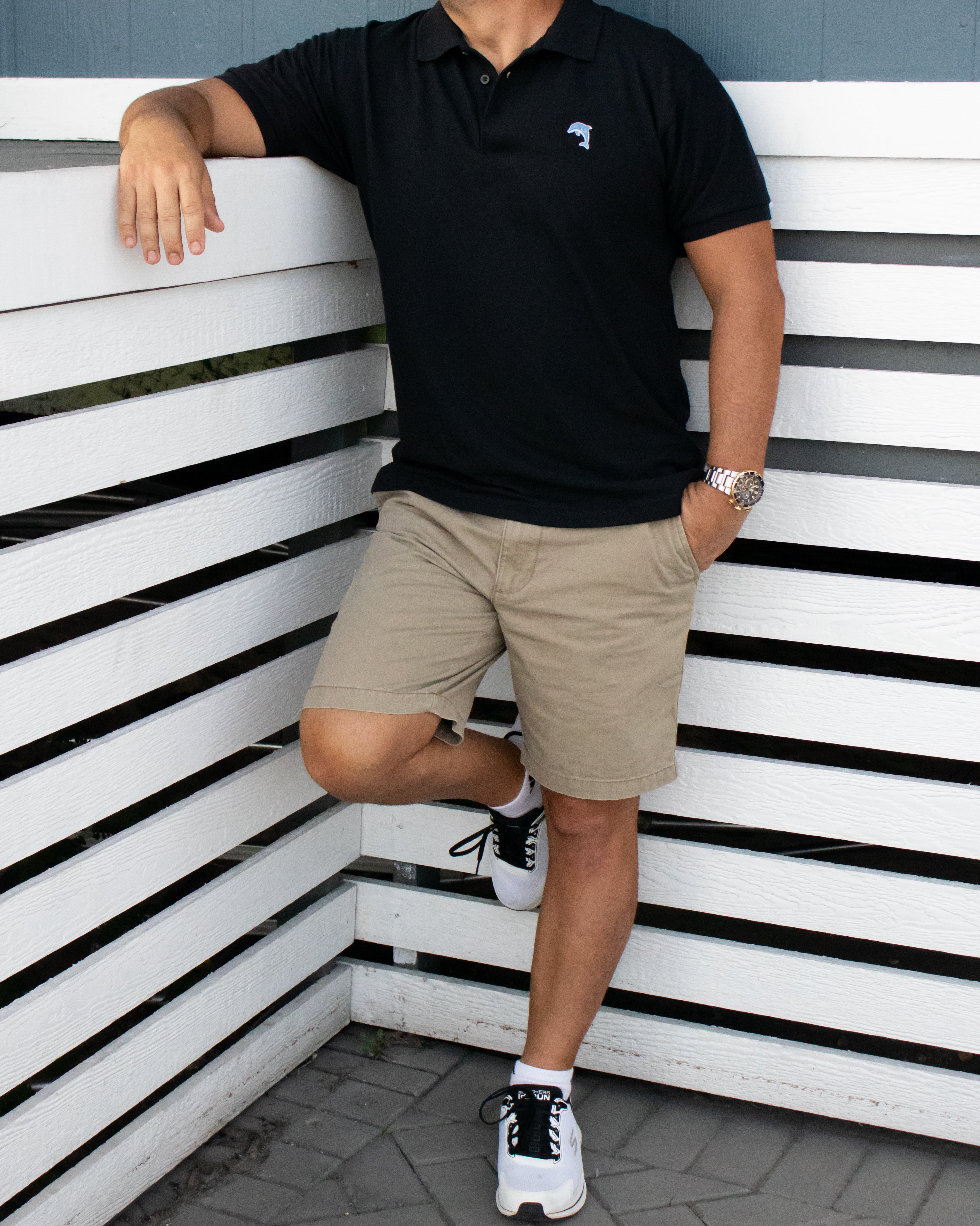 Person wearing a black polo shirt and beige shorts leaning against a white wall with black stripes.