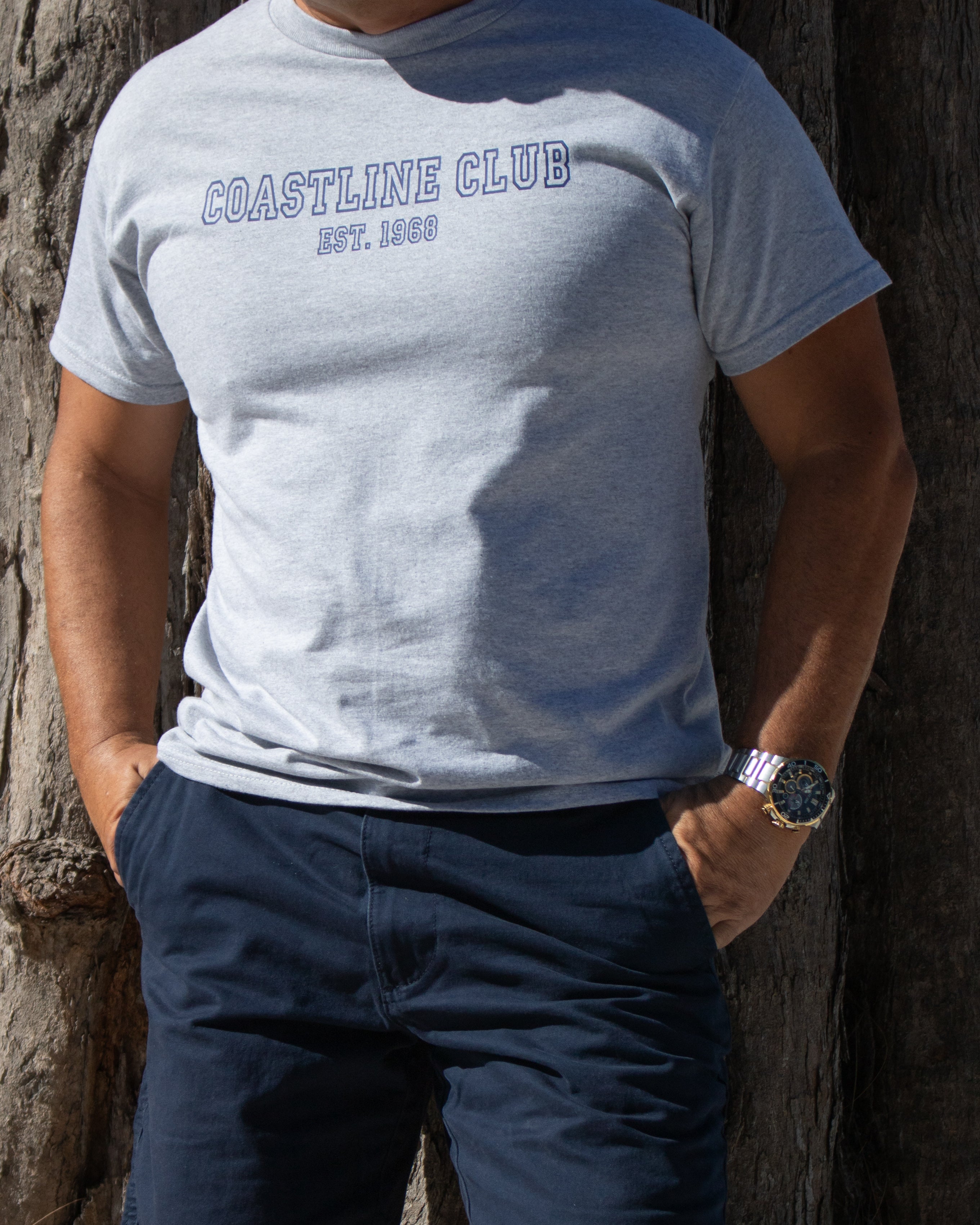 Person wearing a gray 'Coastline Club' t-shirt leaning against a tree.