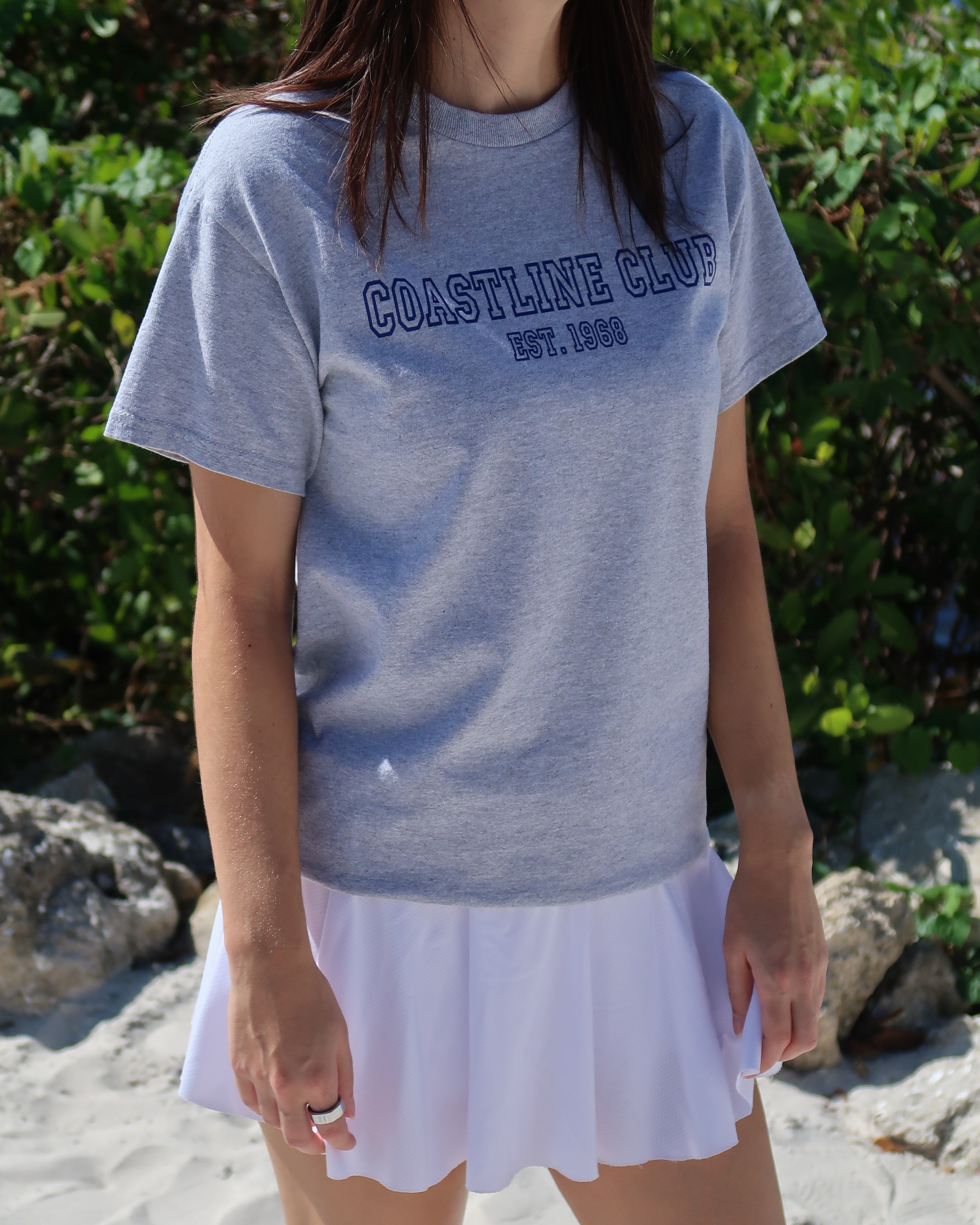 Woman wearing a gray t-shirt with 'Coastline Club' text and a white skirt on a beach.