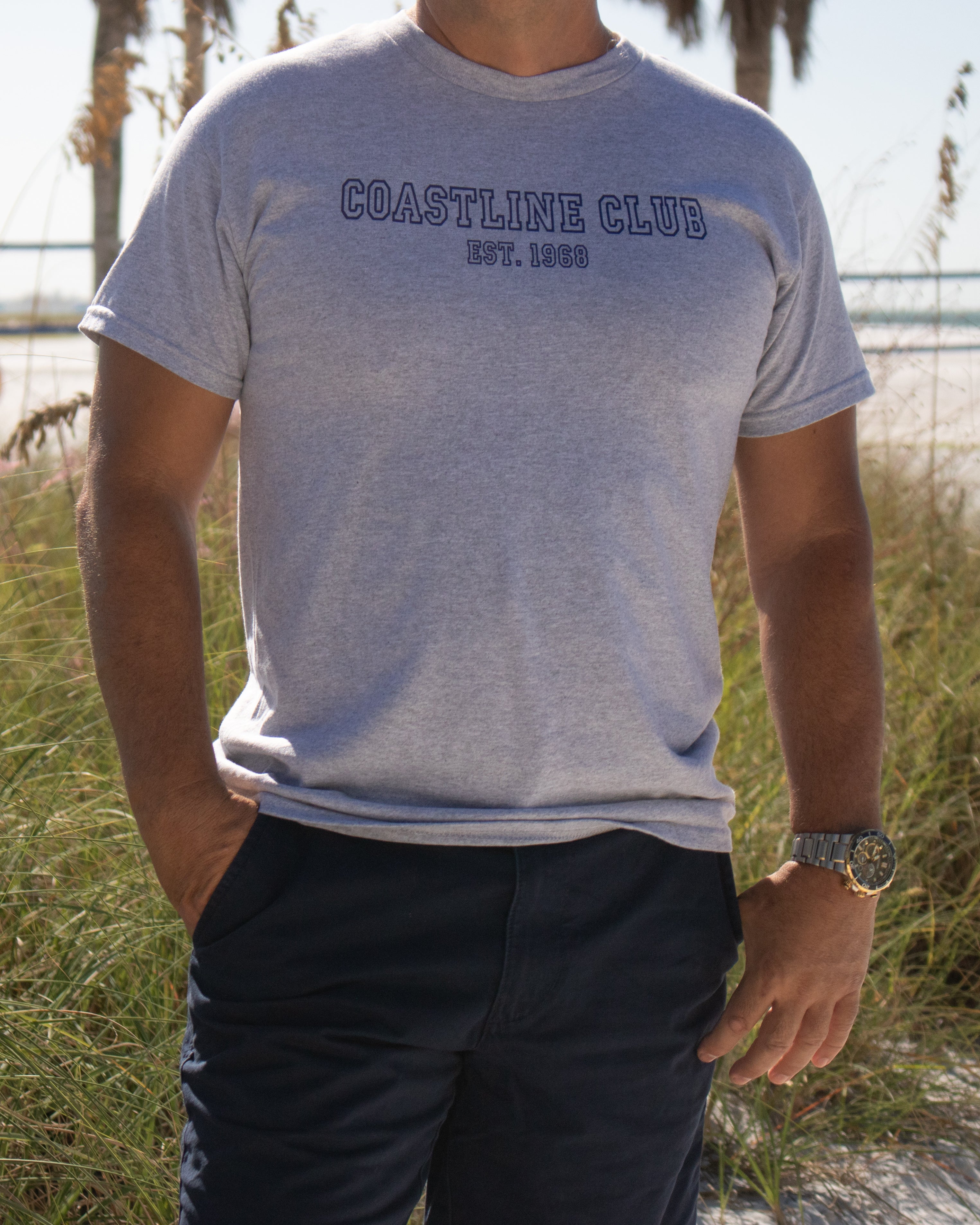 Person wearing a gray 'Coastline Club' t-shirt in a grassy outdoor setting.