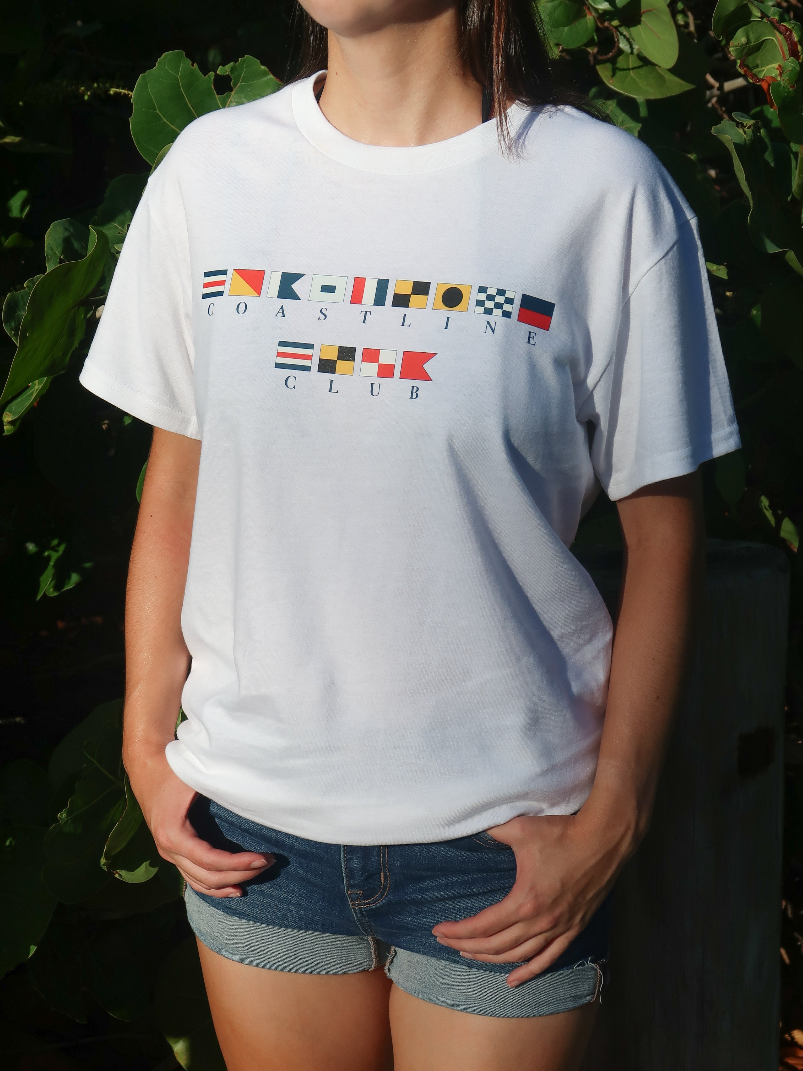 Person wearing a white t-shirt with maritime flag graphic and coastline club text, standing against a dark background with greenery.