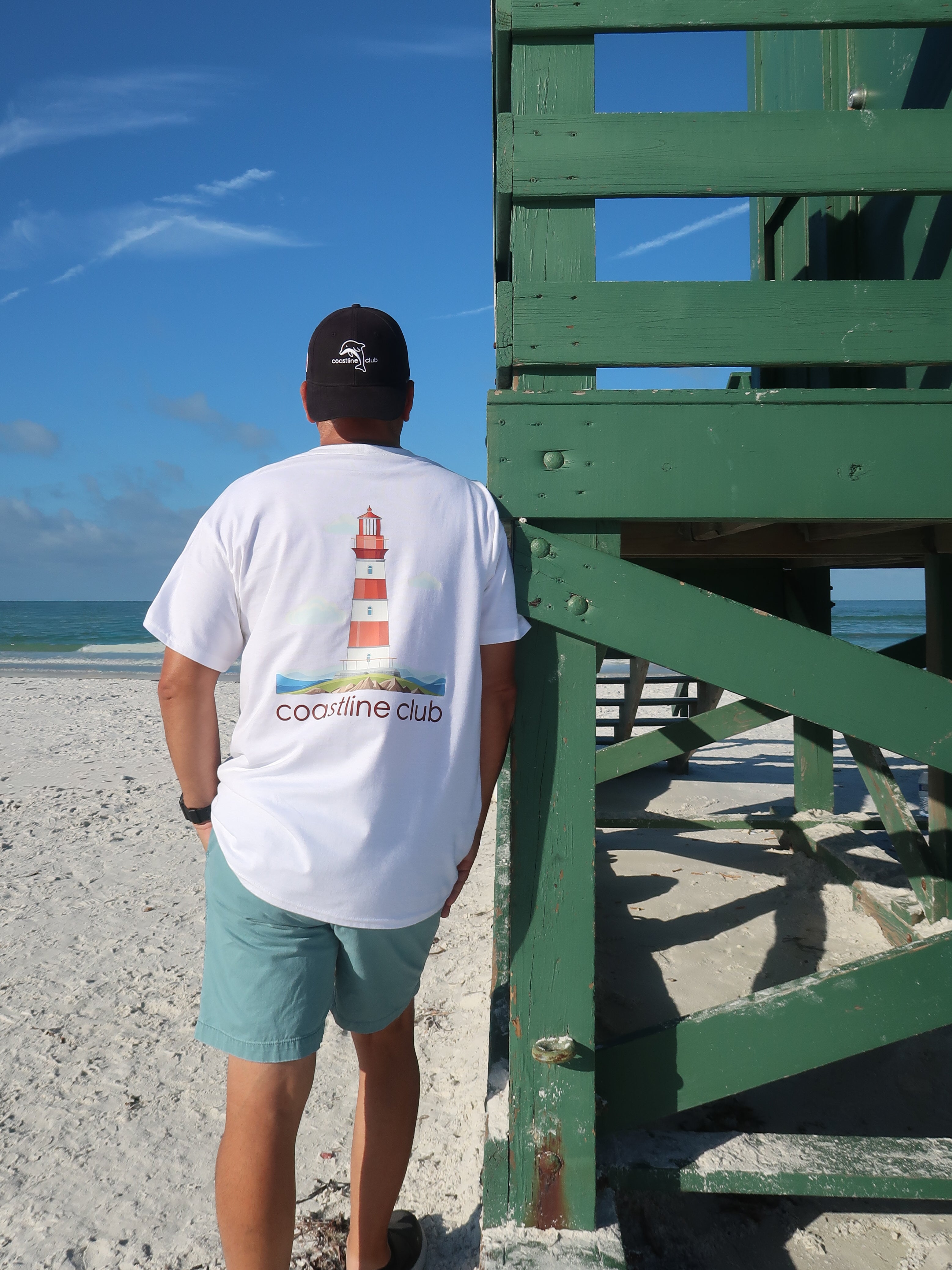 Man wearing a white t-shirt with a lighthouse design and a backwards ball cap on a beach.