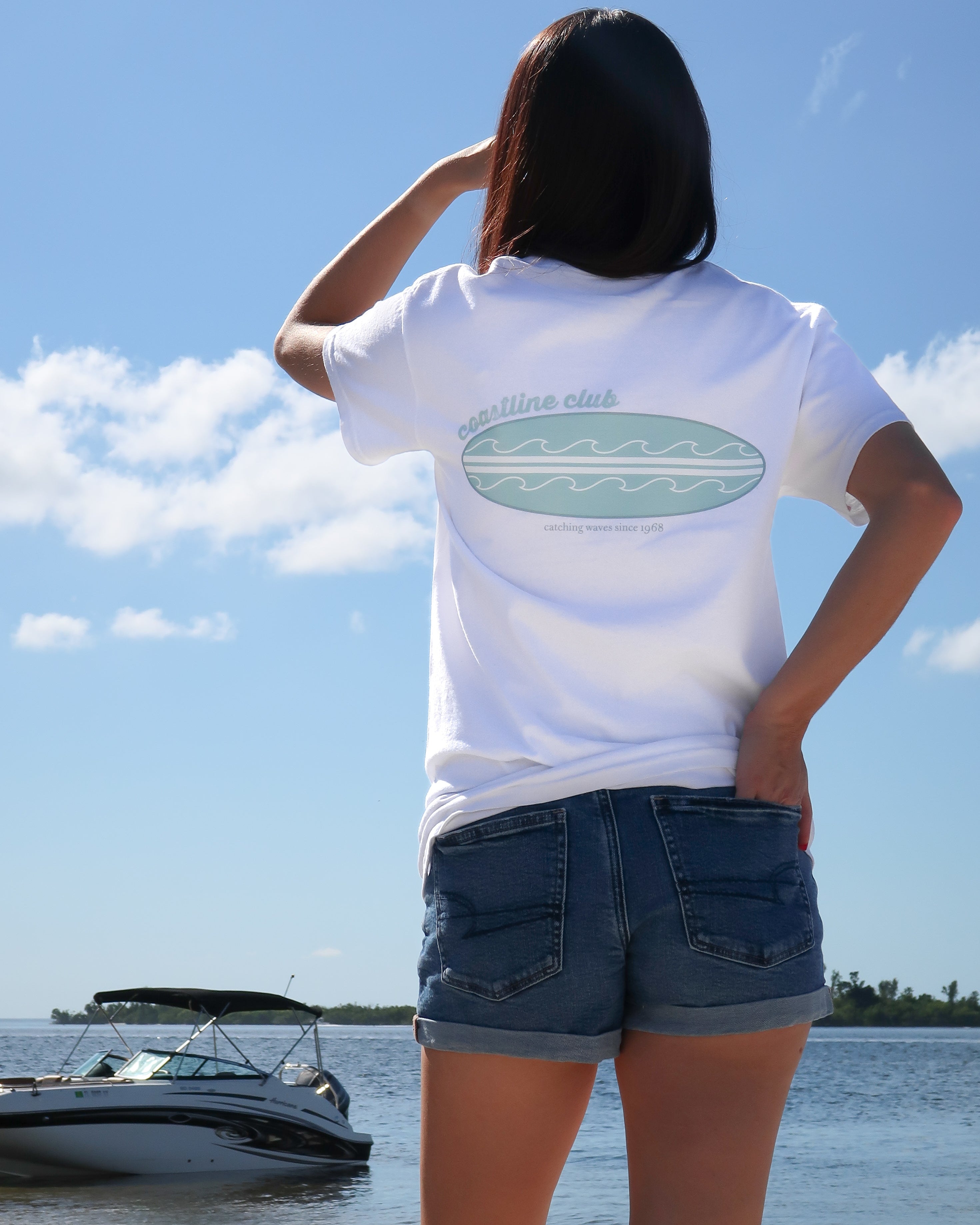 Woman wearing a white t-shirt with a surfboard graphic on the back, standing by a body of water with a boat in the background.