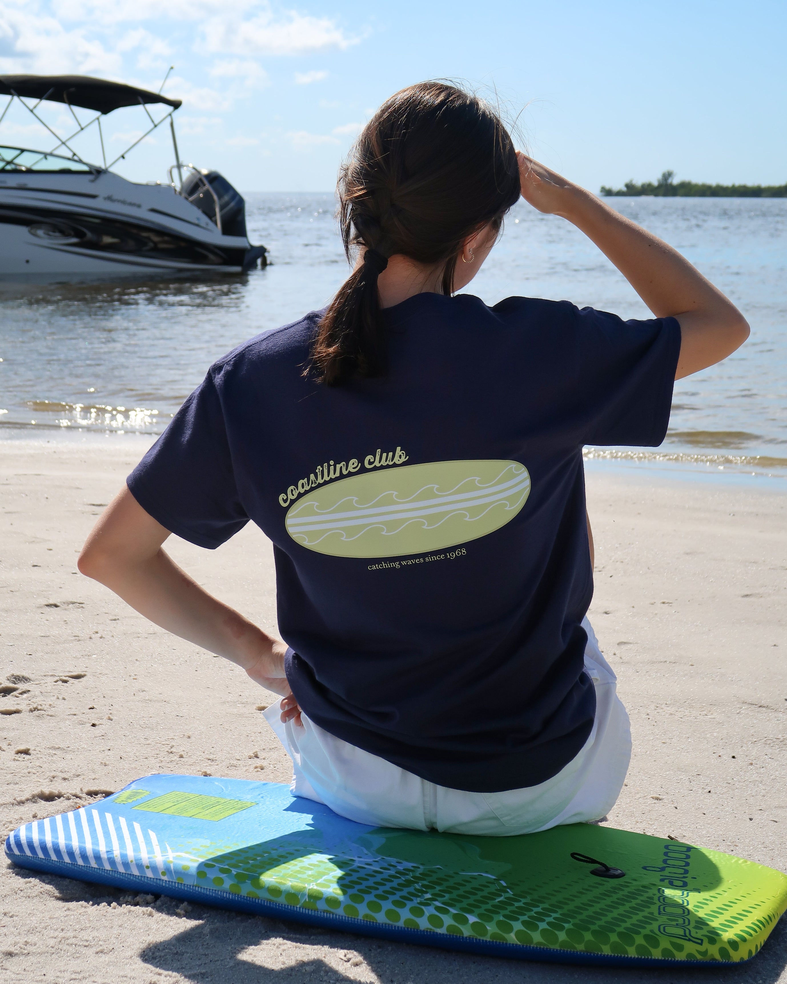 Person sitting on a surfboard with a 'coastline club' t-shirt by a body of water.