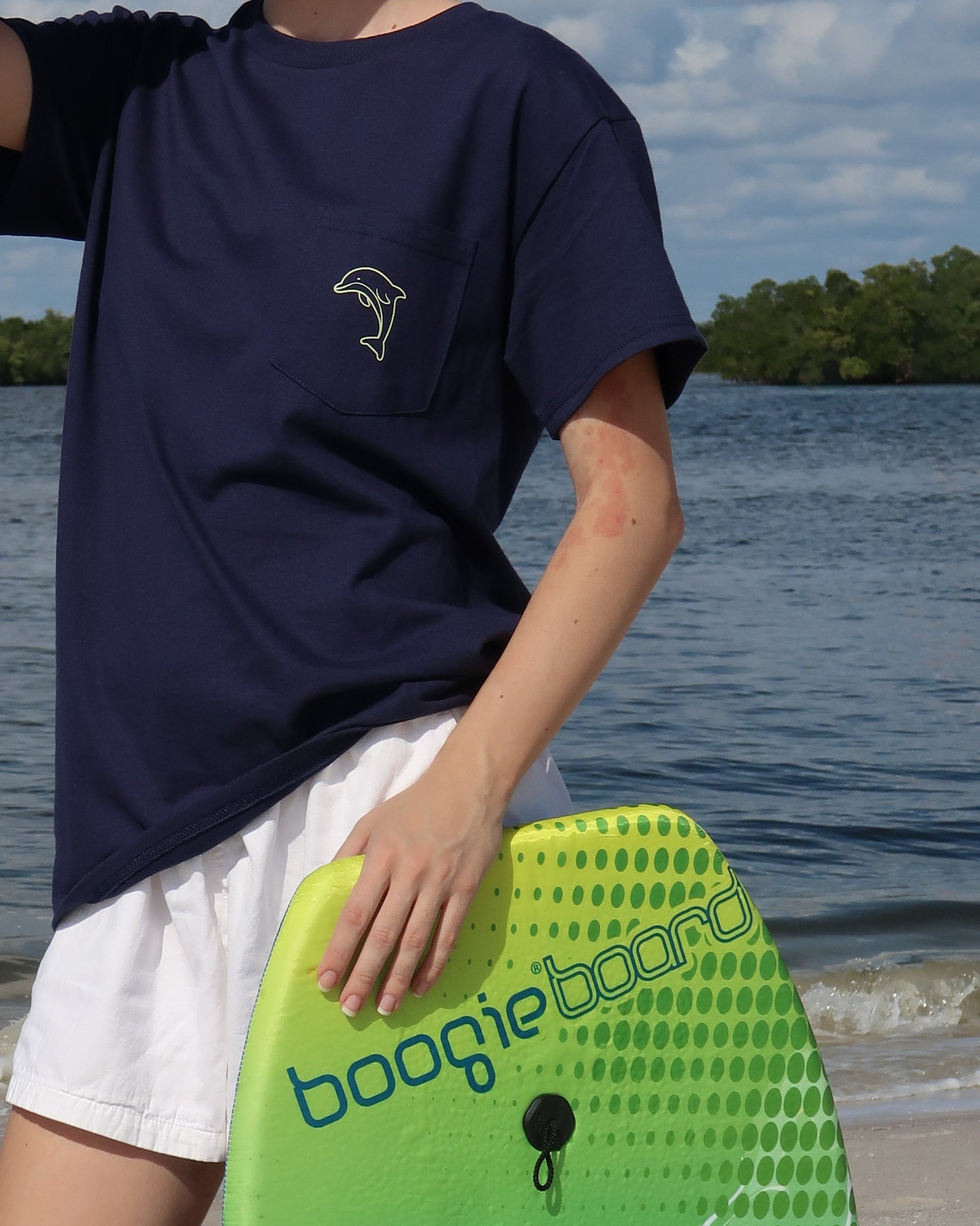 Person holding a green boogie board with 'boogieboard' text, wearing a navy blue shirt and white shorts, standing by a body of water.