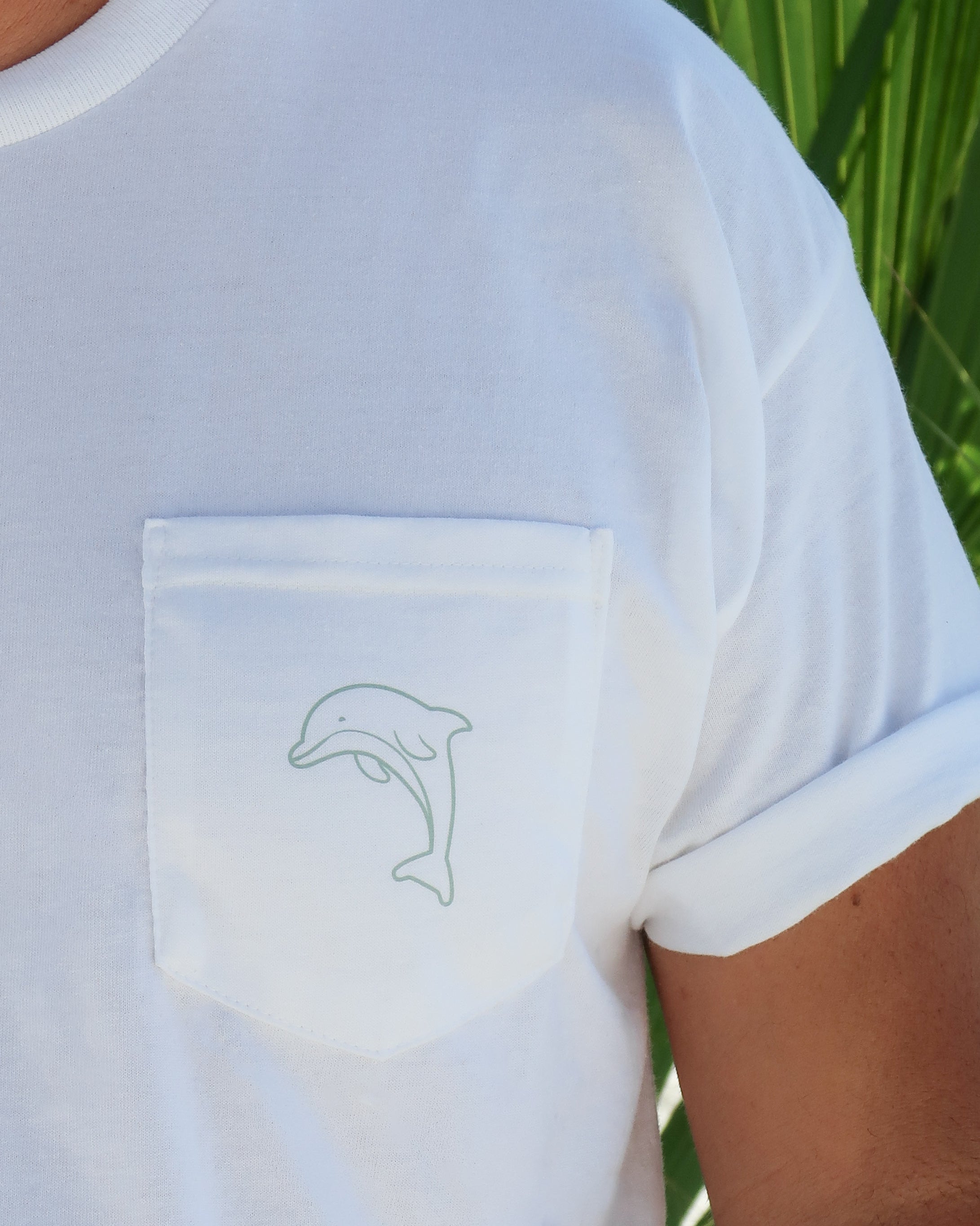 White t-shirt with a dolphin design on the chest pocket against a green leafy background