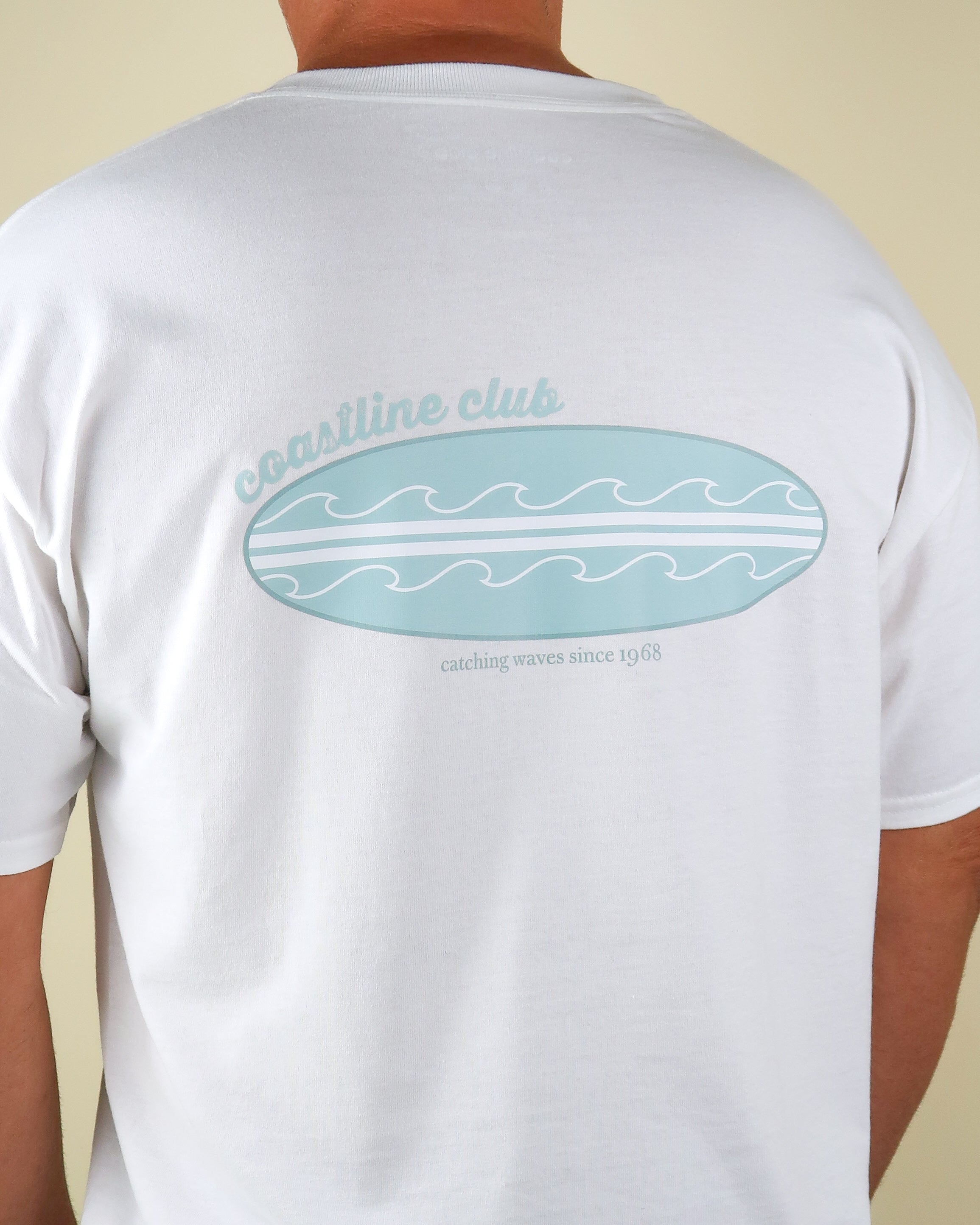 White t-shirt with a blue surfboard graphic and coastline club text on the back, worn by a person.