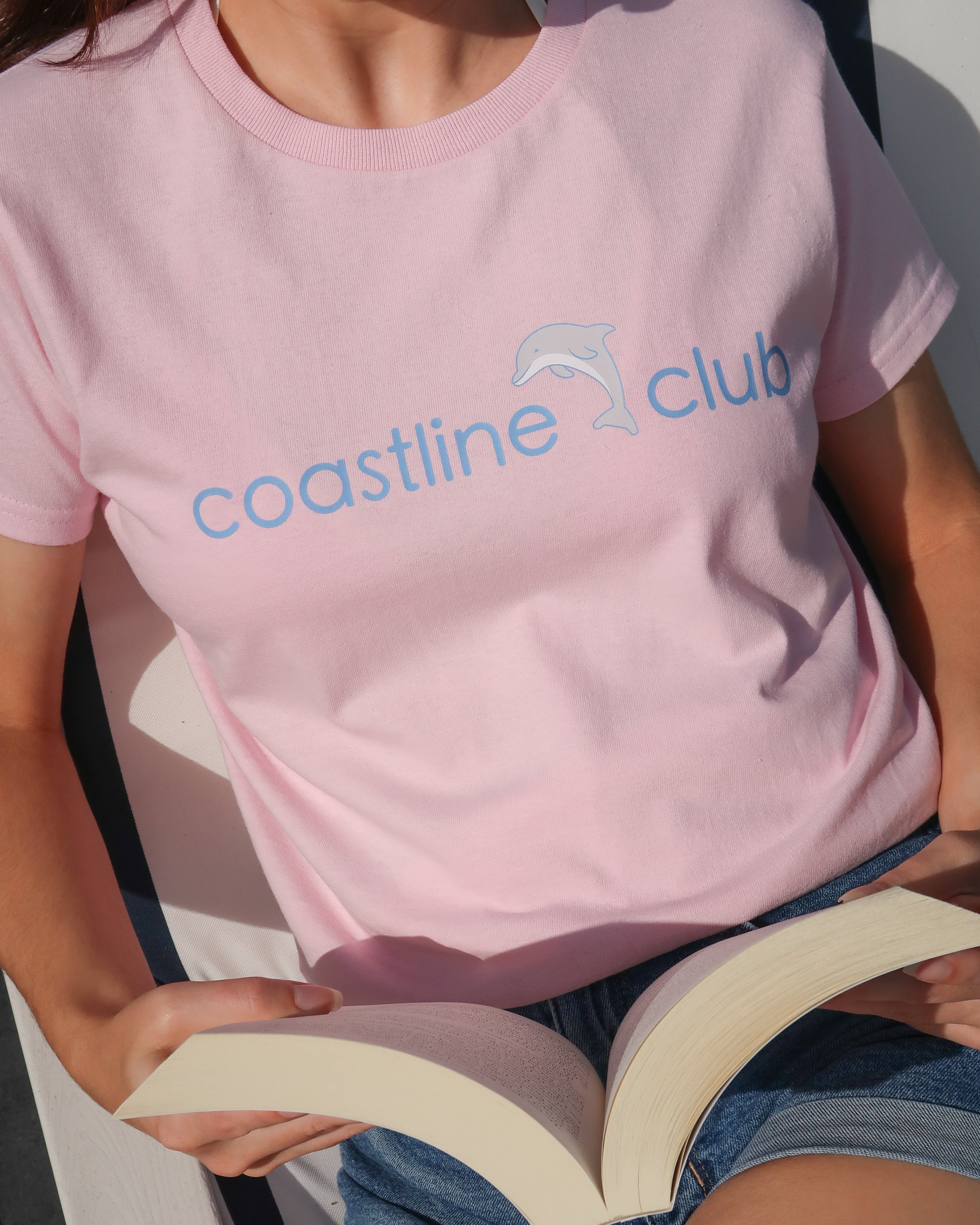 Person wearing a pink 'coastline club' t-shirt holding an open book.