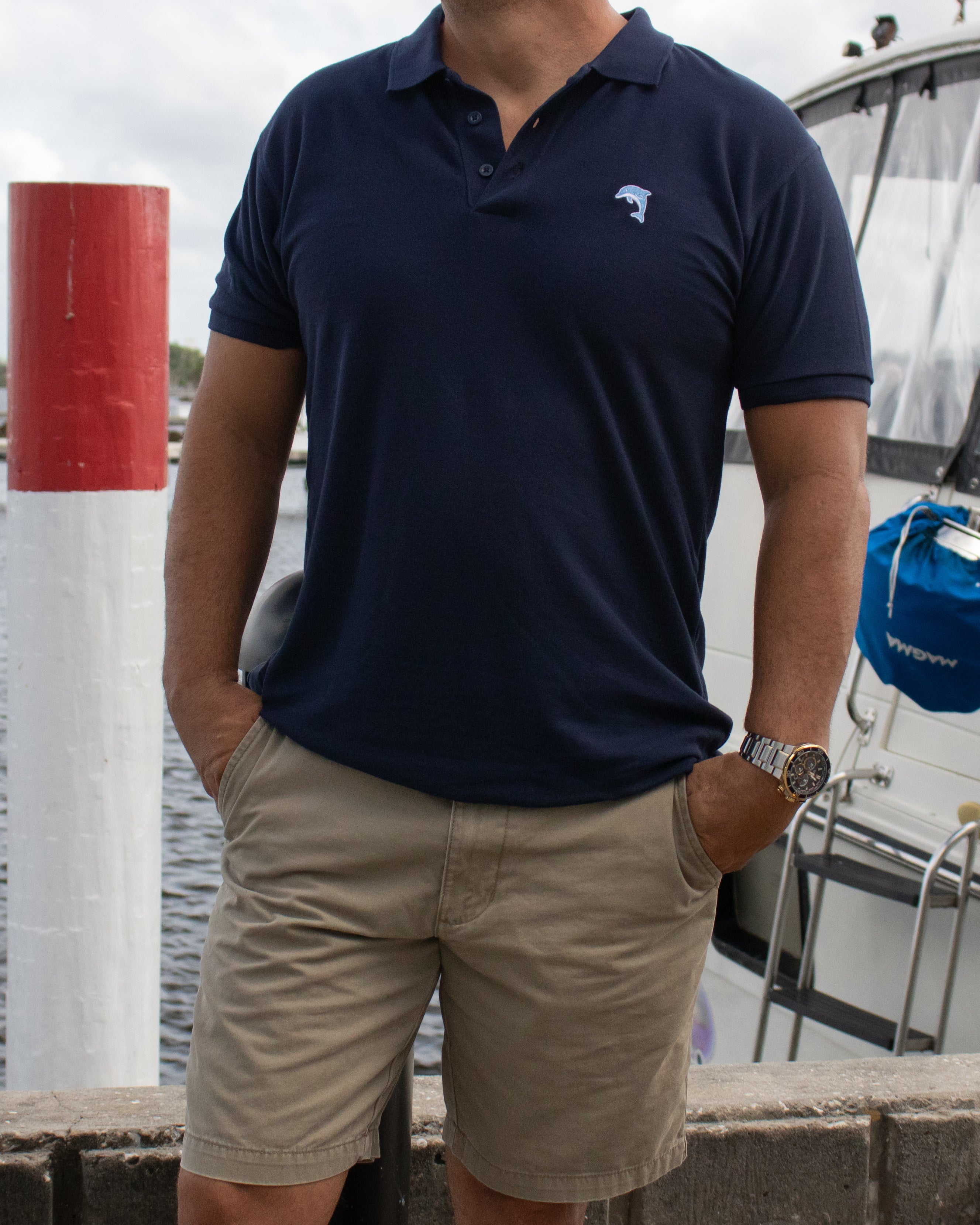 Person wearing a navy polo shirt with a dolphin logo and beige shorts by a boat