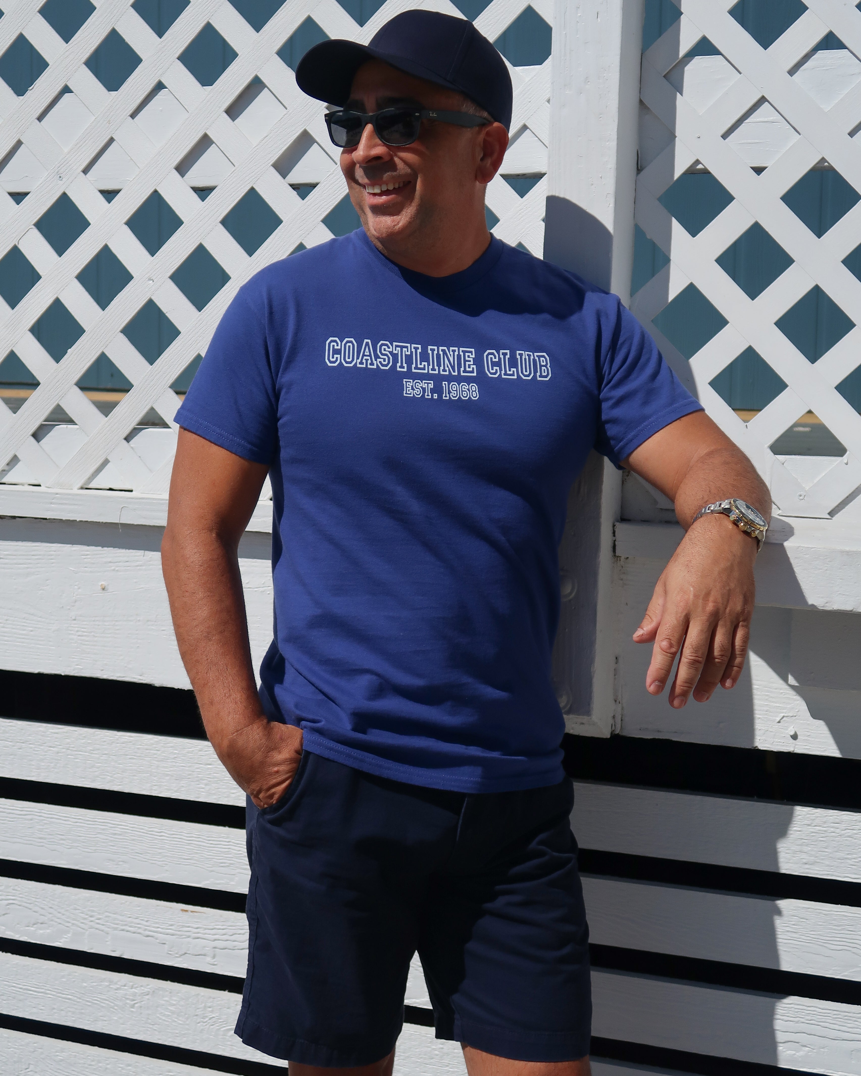 Man wearing a blue 'Coastline Club' t-shirt and navy shorts standing against a white lattice background.