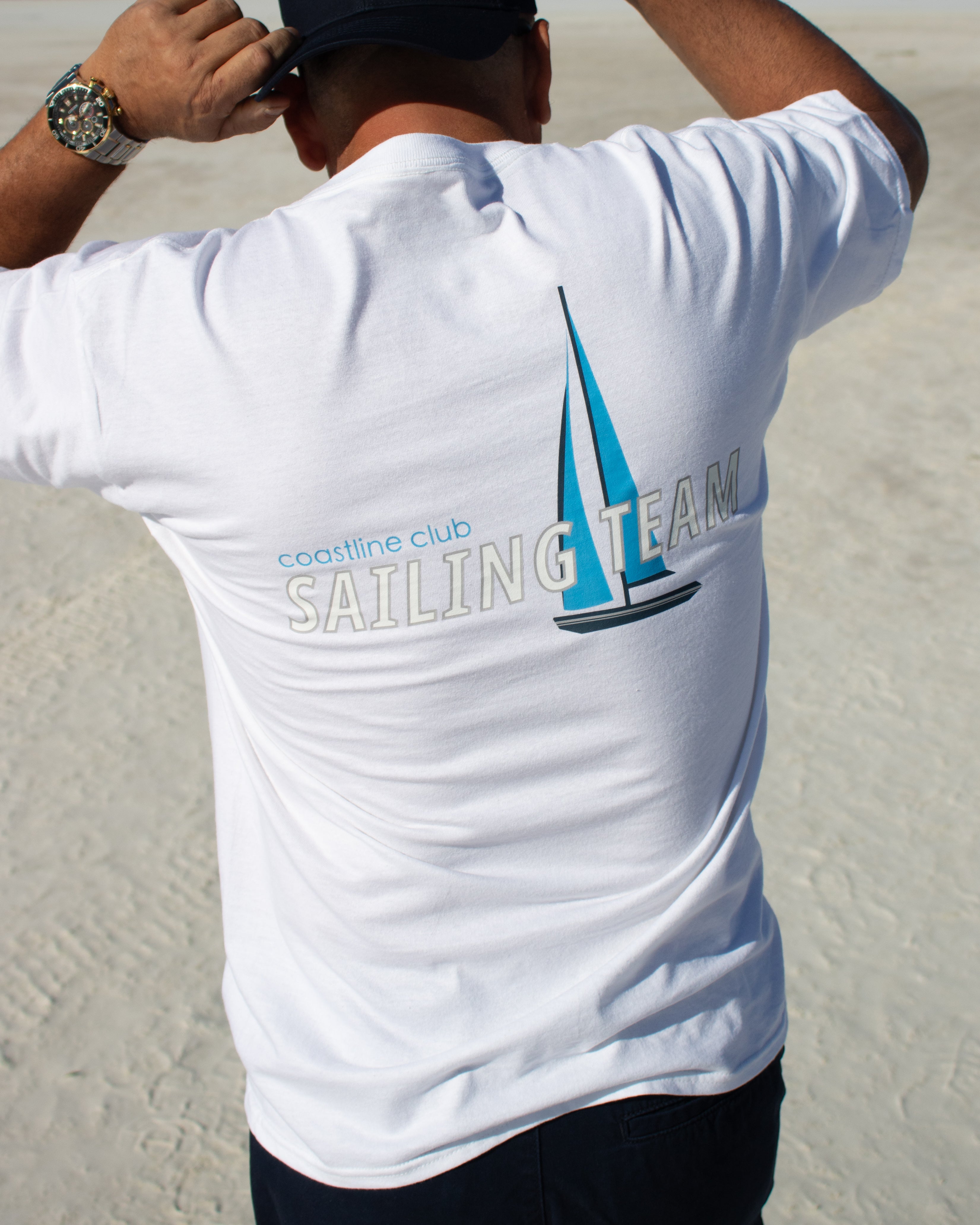 Person wearing a white t-shirt with 'coastline club SAILING TEAM' text and logo on a sandy background