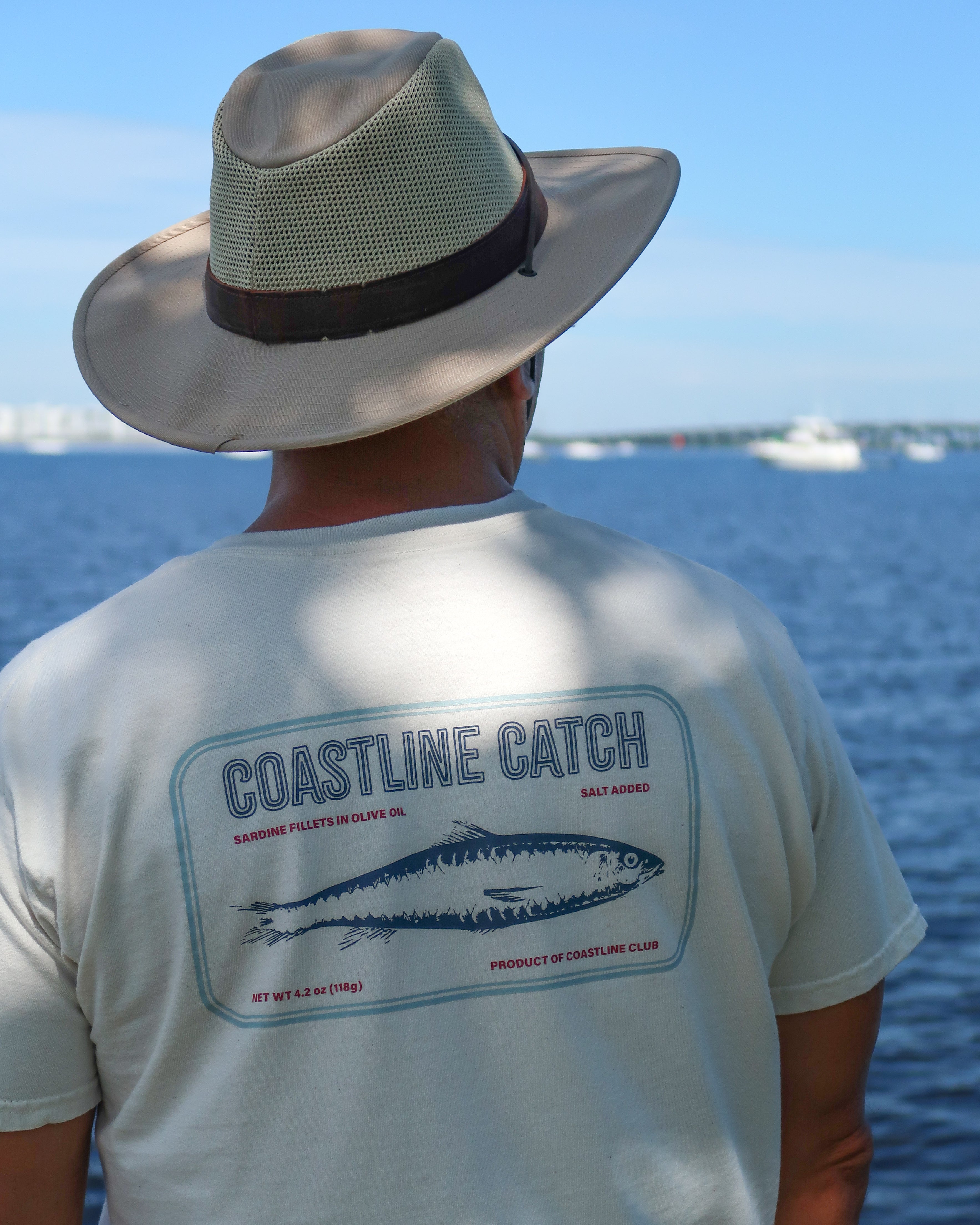 Person wearing a 'Coastline Catch' sardine t-shirt by the water