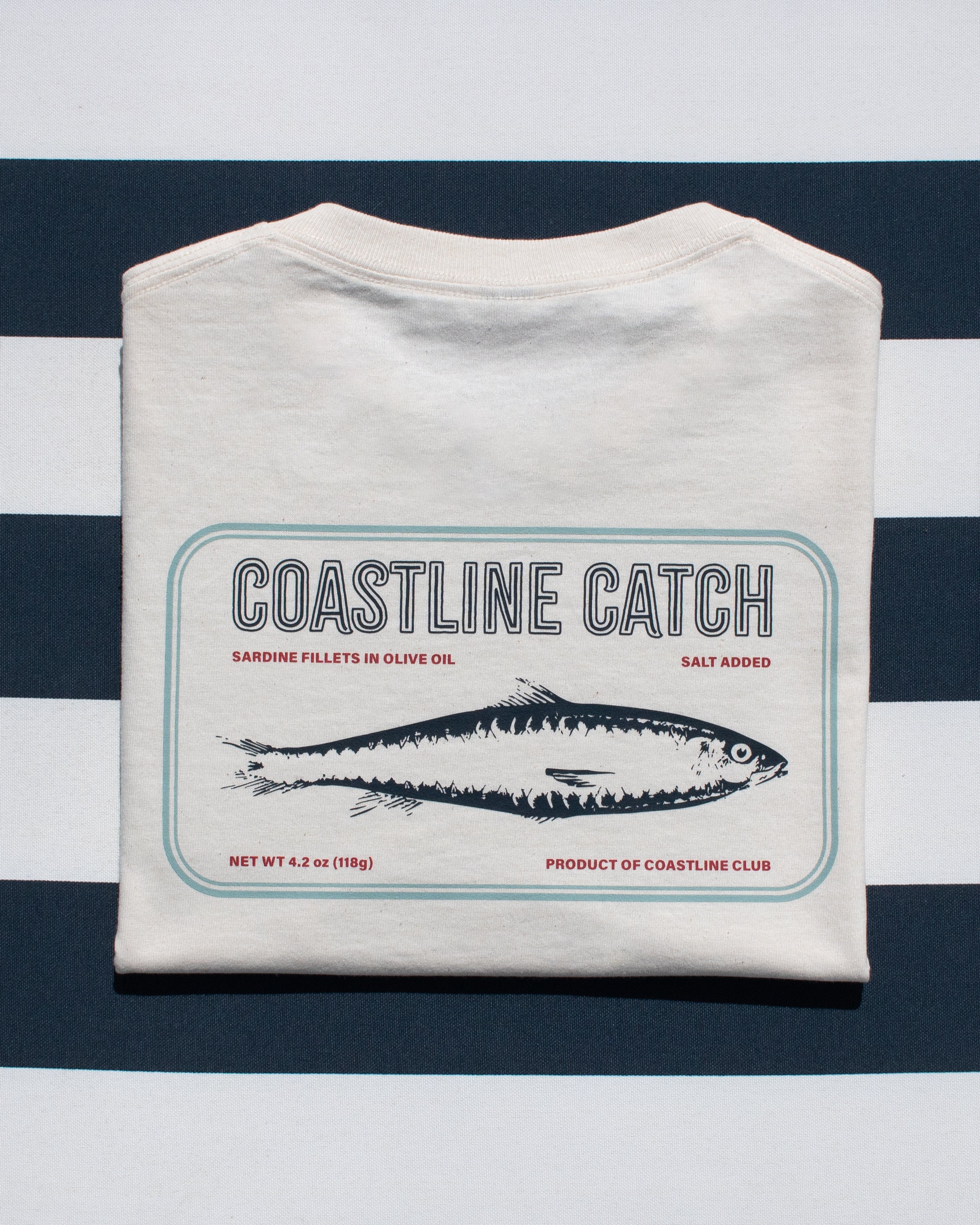 Folded white t-shirt with 'Coastline Catch' sardine can label on a blue and white striped background
