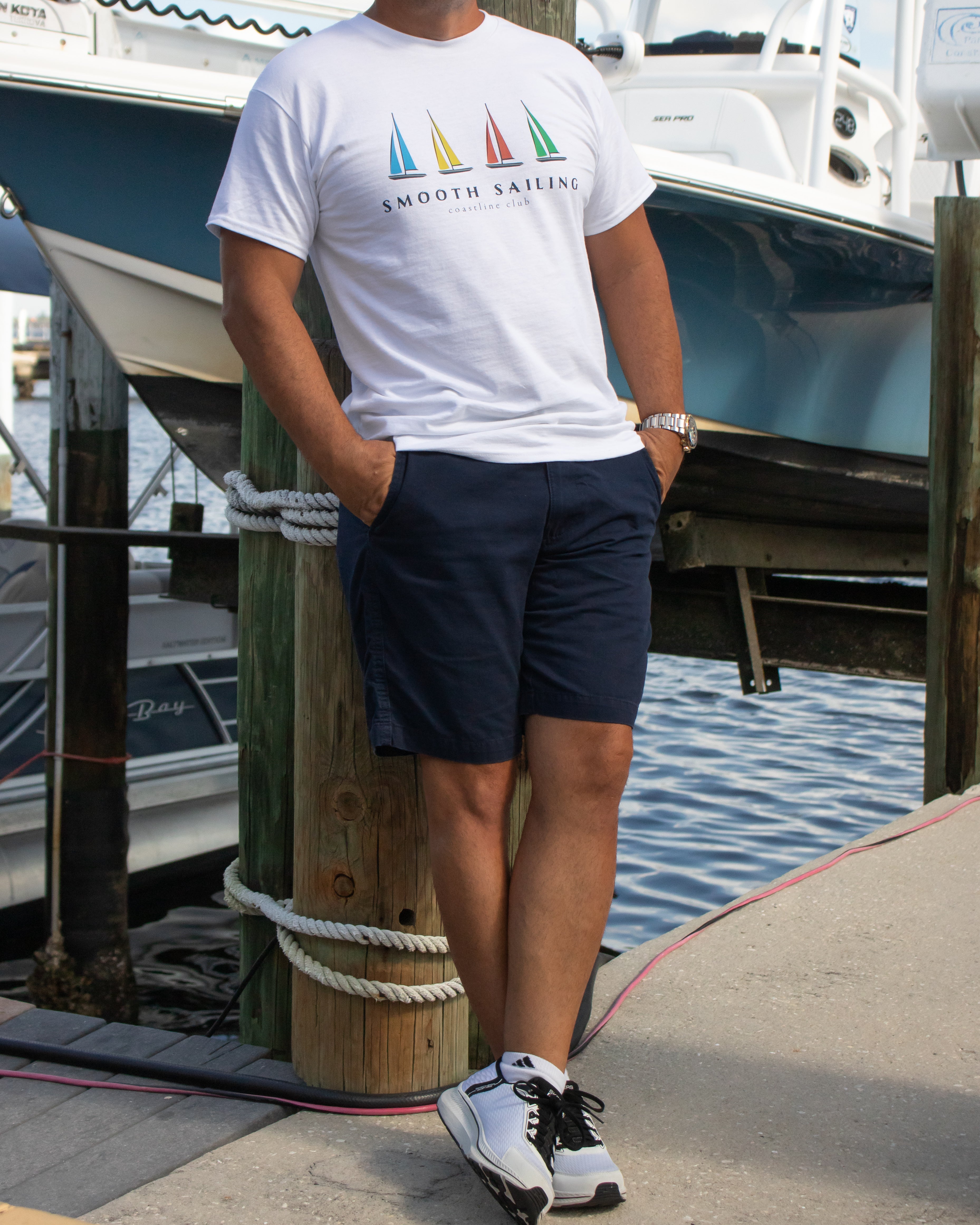 Person wearing a white t-shirt with a sailboat design and text, standing on a dock with boats in the background.