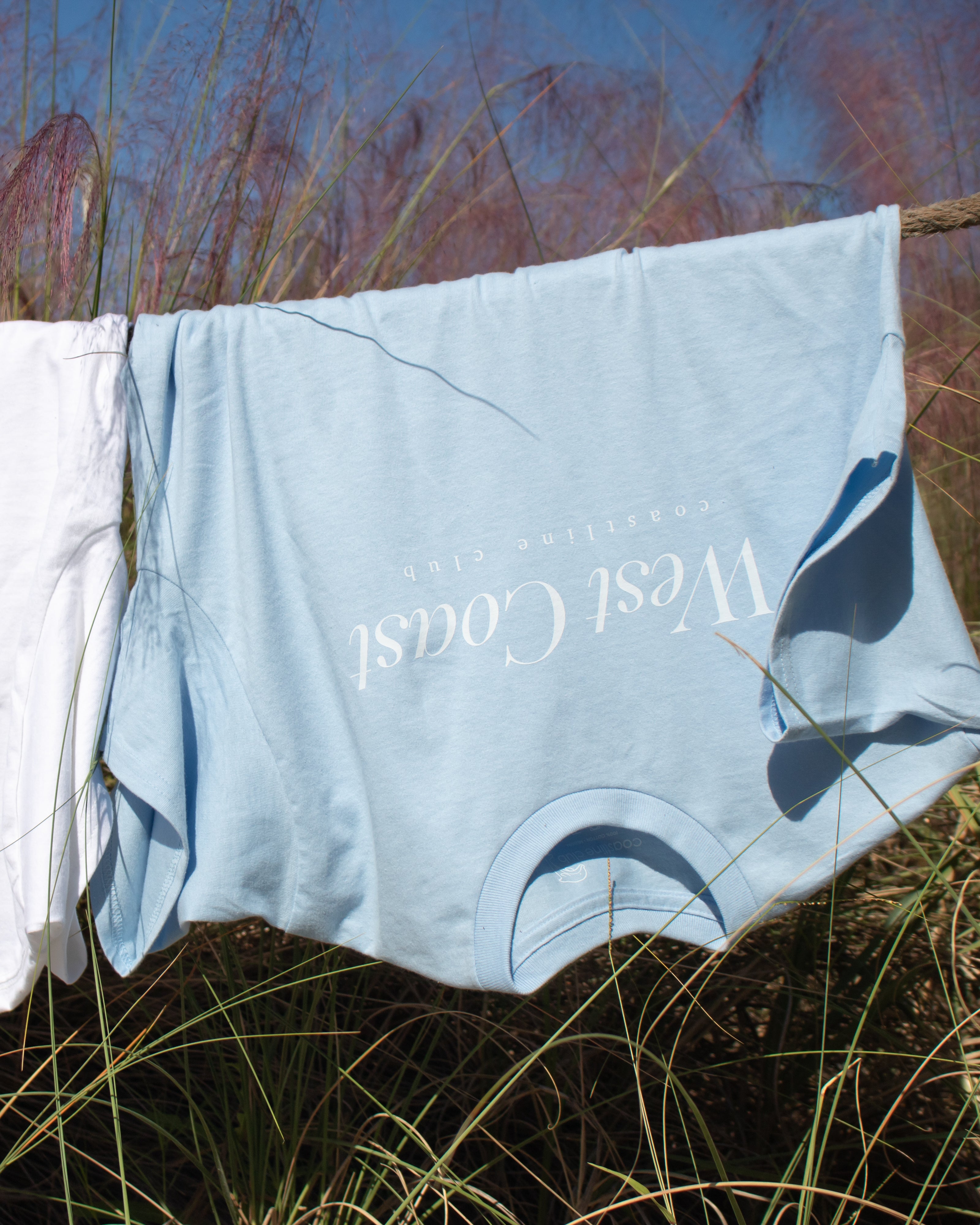Light blue shorts with 'West Coast' branding hanging on a clothesline against a natural background.