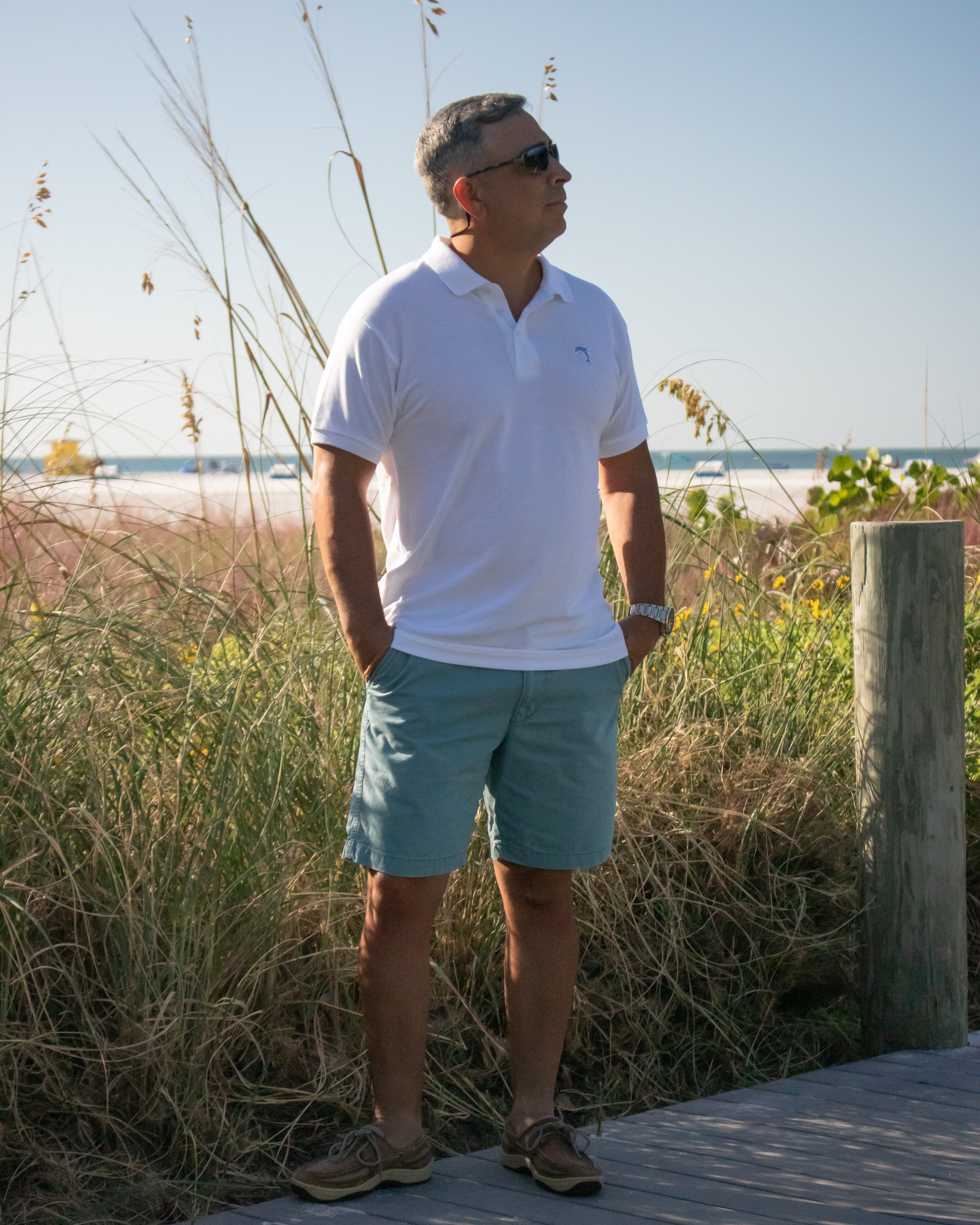 Man standing on a wooden boardwalk by a beach with tall grasses and a clear sky.