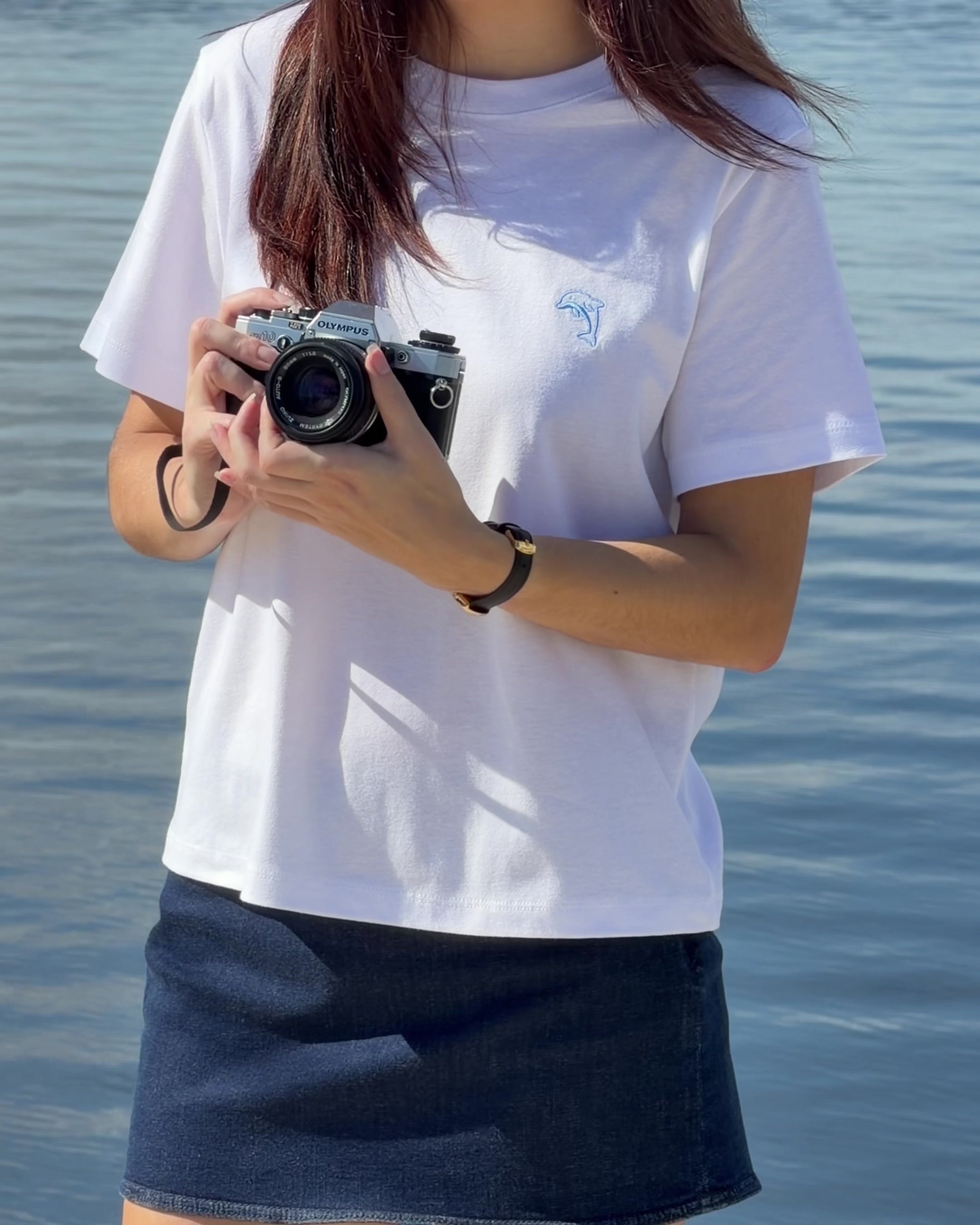 person wearing white shirt with blue dolphin logo and denim skirt holding a film camera