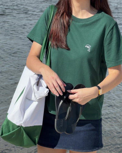 person wearing a green shirt with a white dolphin logo holding black sandals and a white and green tote bag