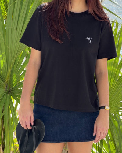 person wearing a black shirt with a white dolphin logo and denim skirt holding black sandals