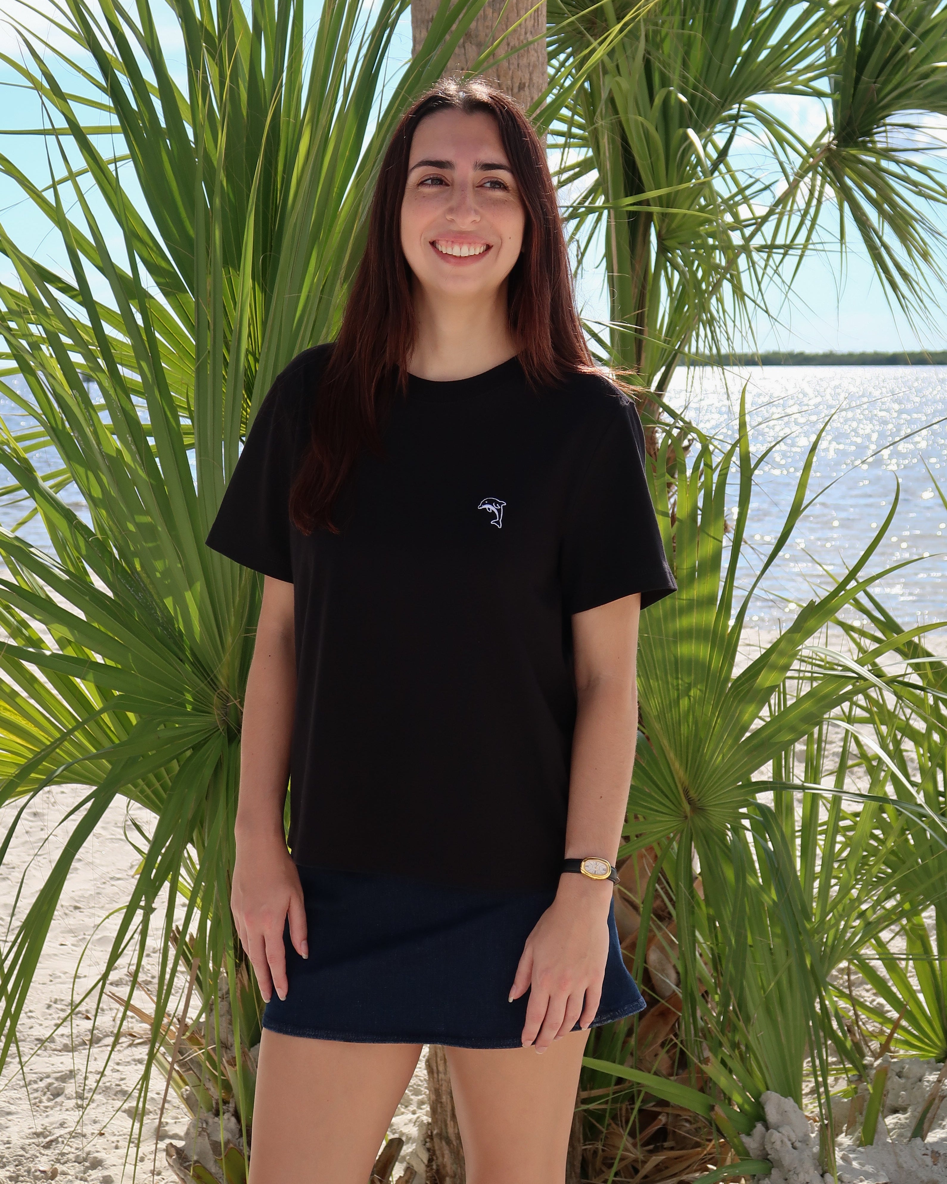 Woman in a black t-shirt and navy skirt standing in front of palm trees and water.