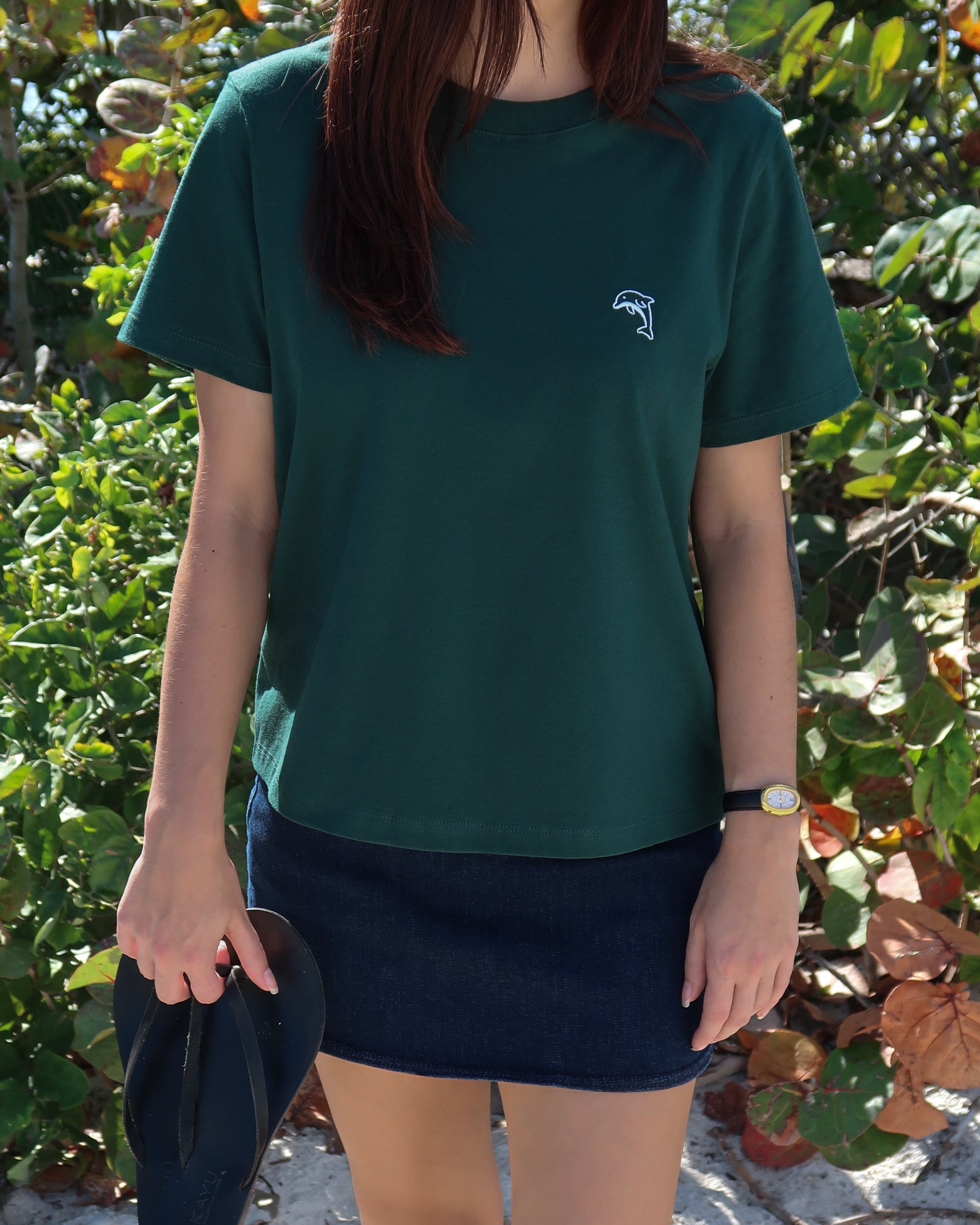 Person wearing a green t-shirt with a dolphin logo and a denim skirt with sandals in their hand.