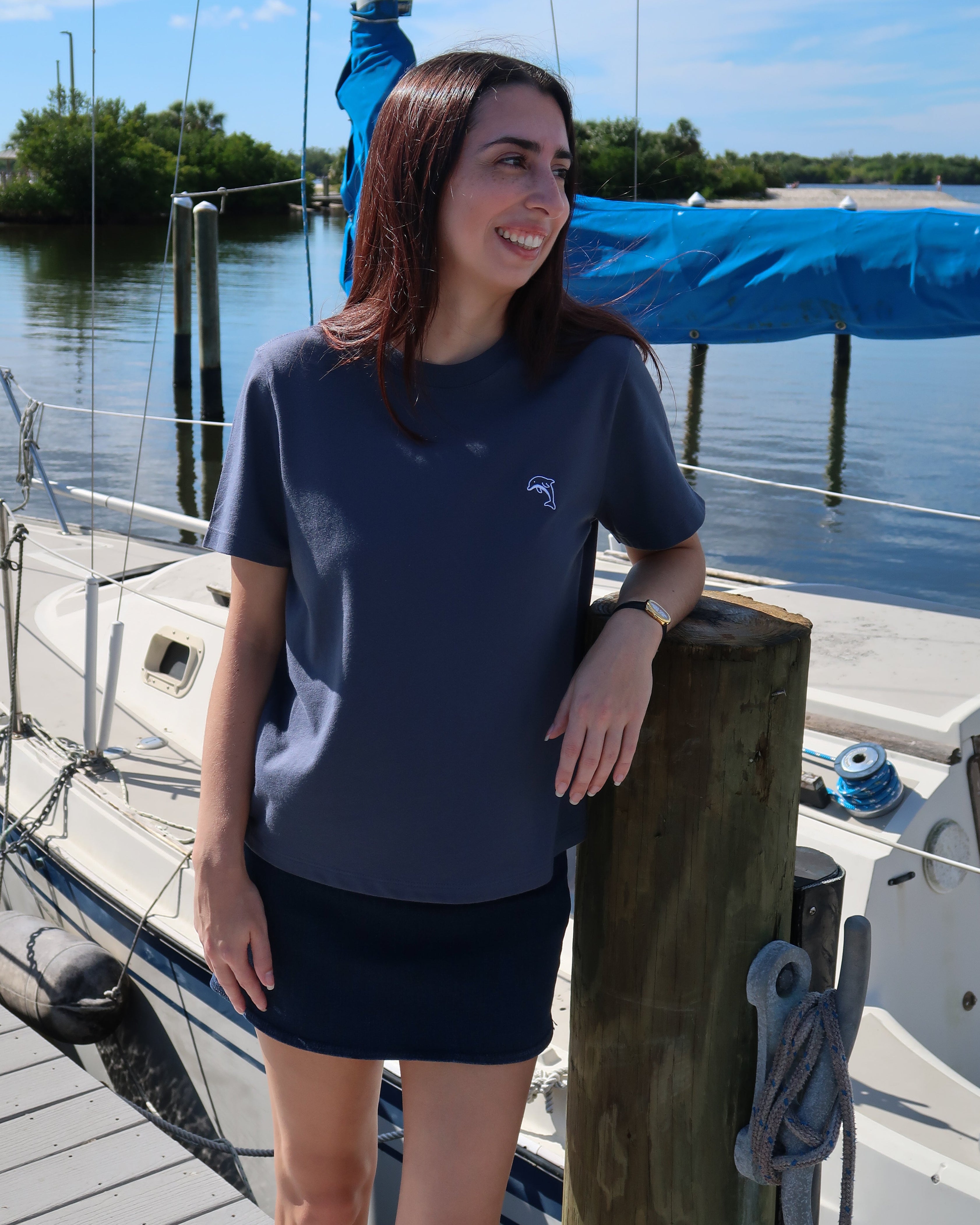 Woman in a steel blue shirt with a dolphin standing on a dock by a sailboat