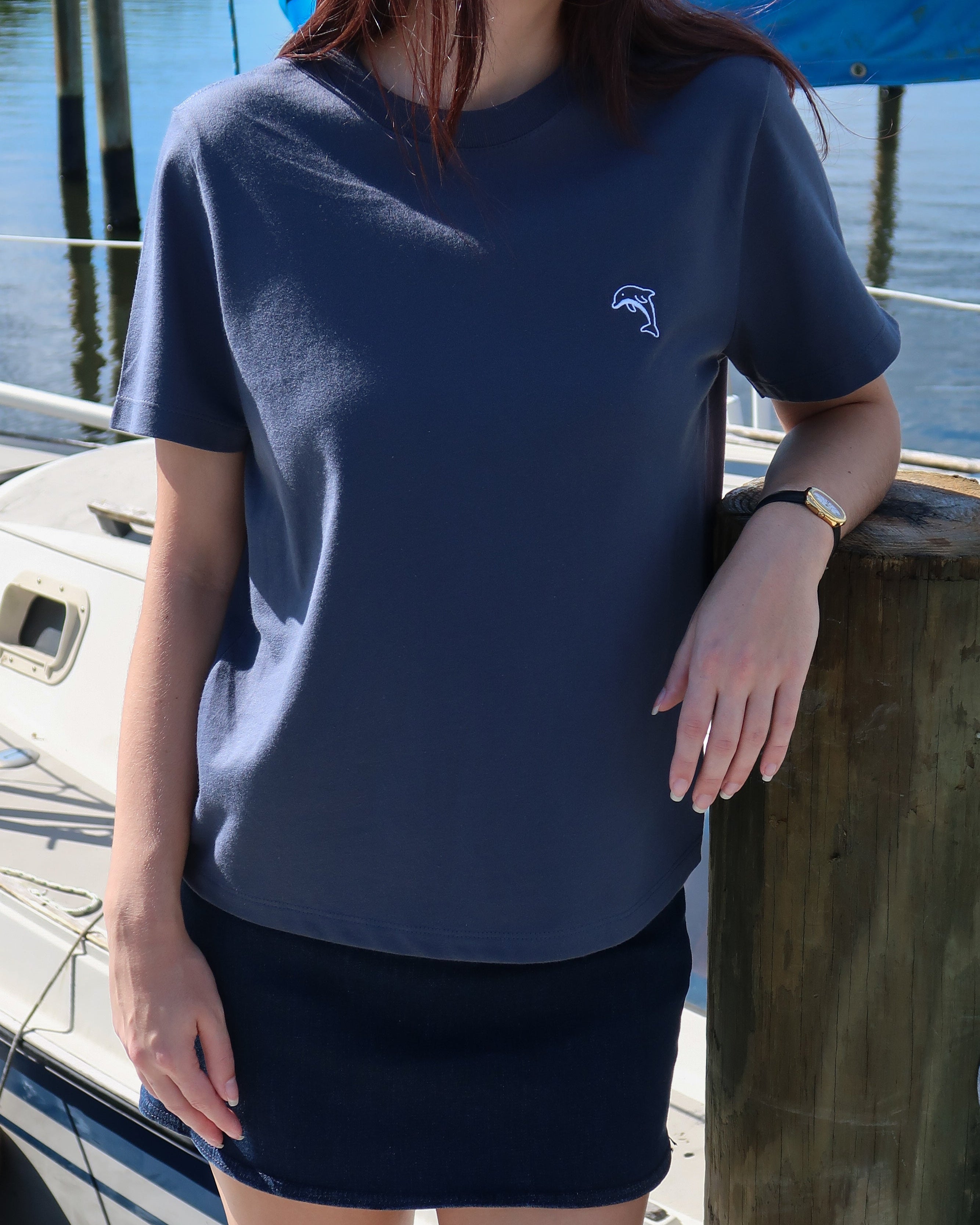 Person wearing a steel blue t-shirt with a dolphin logo, standing by a boat on a dock.