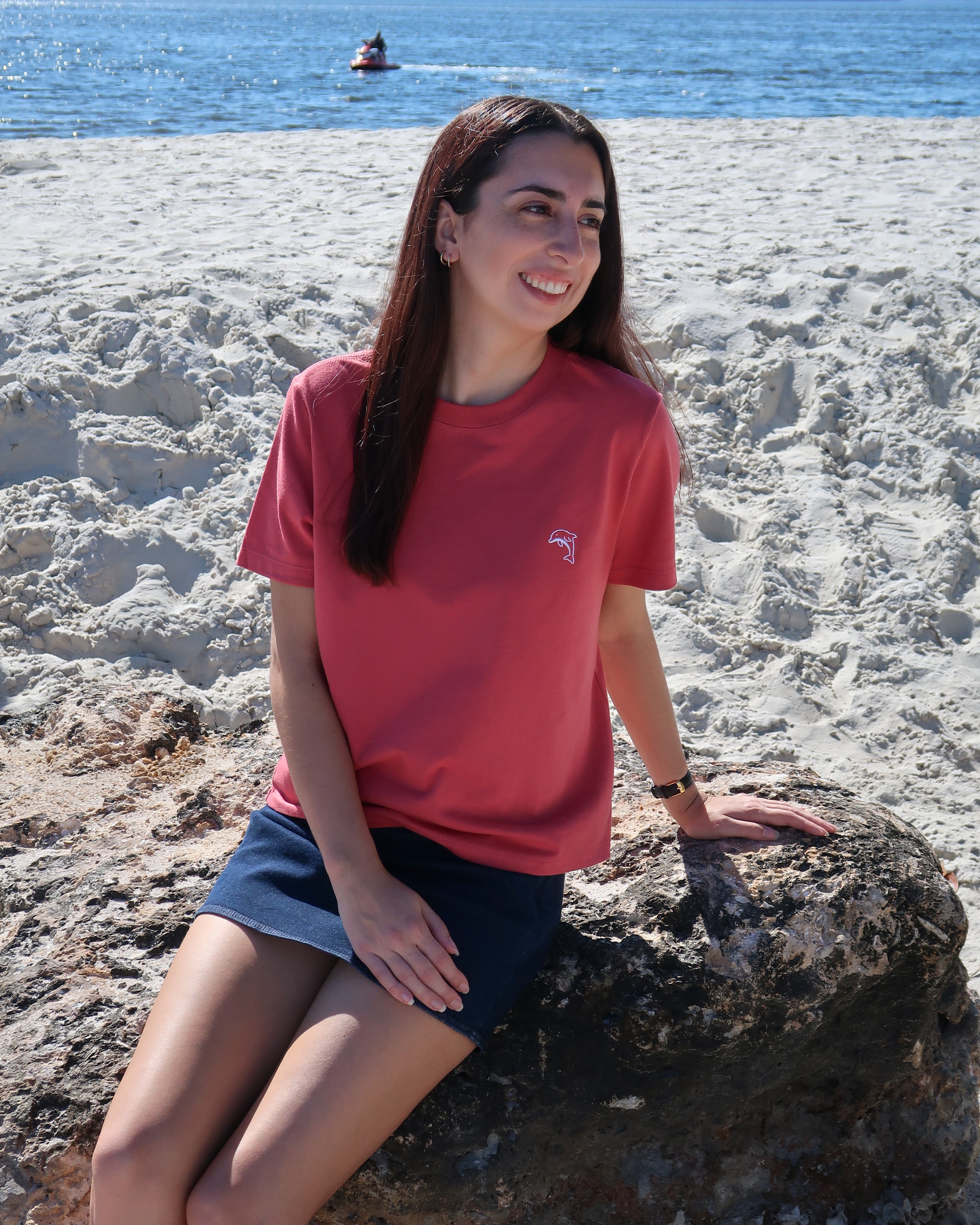 Woman in a red shirt sitting on a rock by the beach with a jetski in the background.